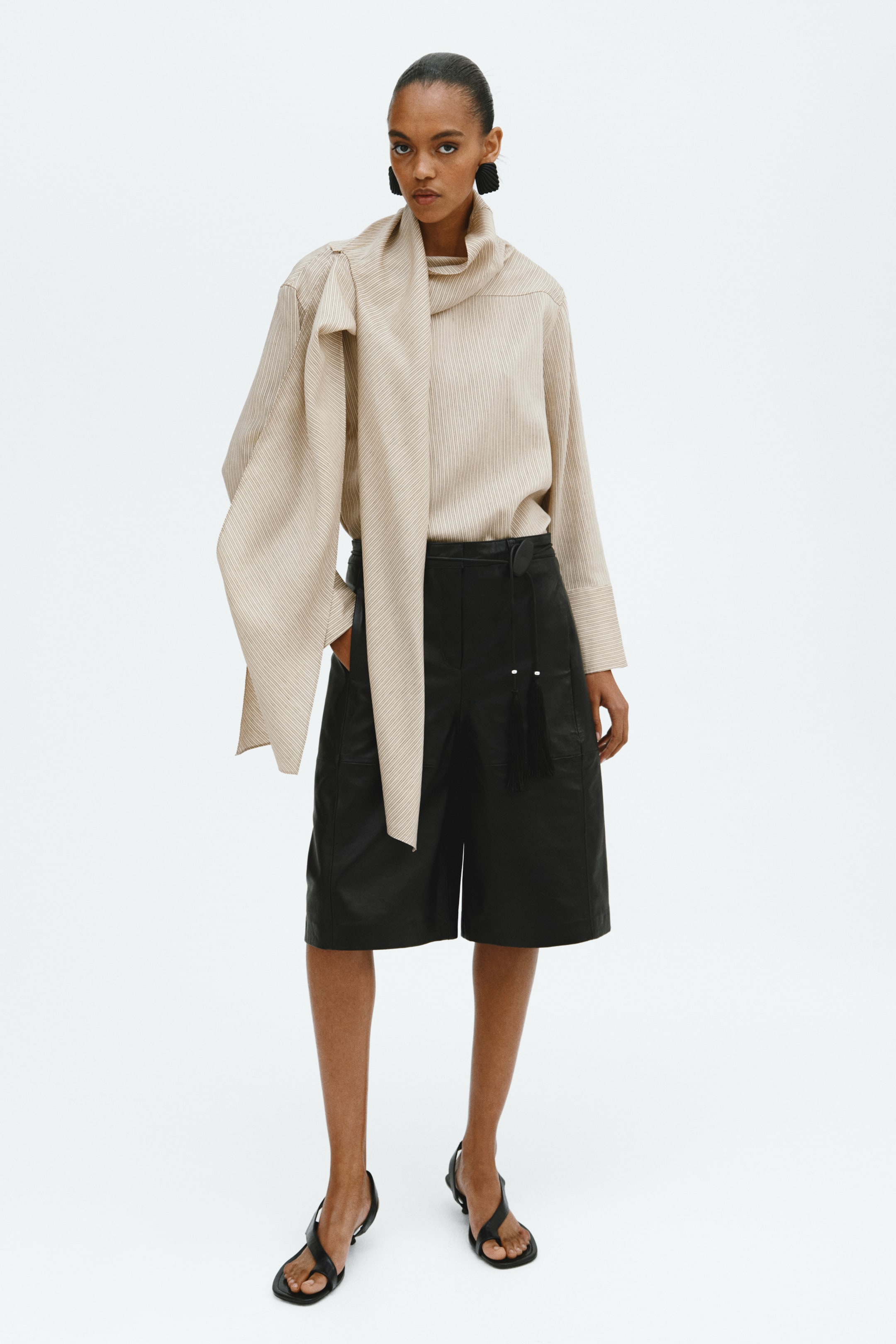 Grotere afbeelding bekijken: Een beige gestreept shirt met lange, wijde mouwen en een geïntegreerde sjaaldetail die rond de hals is gedrapeerd en naar voren loopt. Het wordt gestyled met zwarte, knielange shorts met een leren look, voorzien van een strikriem met kwastjes, en zwarte teenslippers.