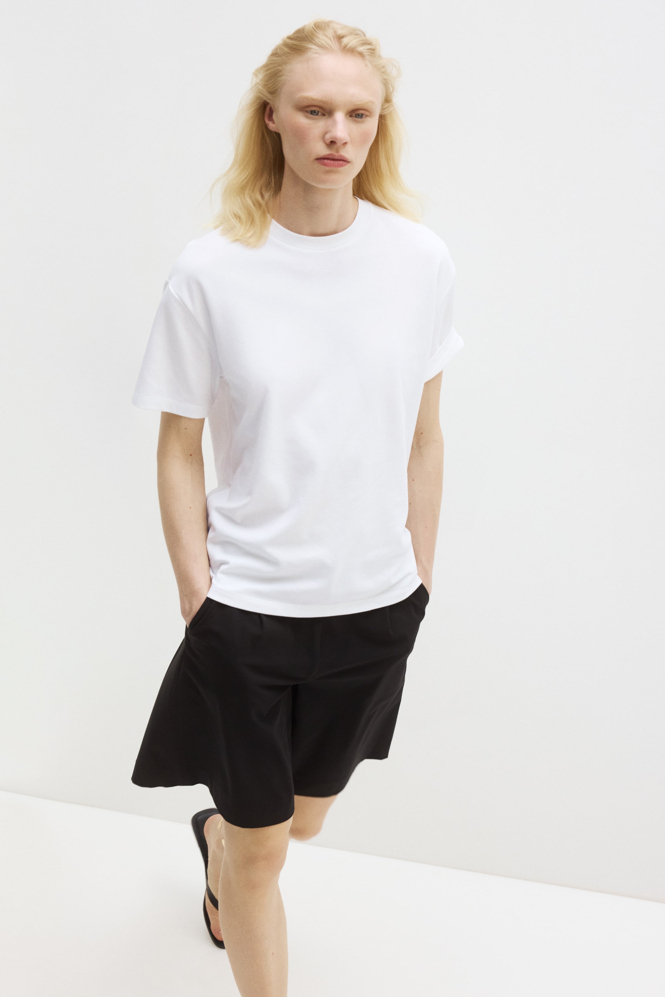 Visualizza immagine più grande: Una t-shirt bianca a girocollo con maniche corte arrotolate e una vestibilità rilassata è abbinata a pantaloncini neri al ginocchio con tasche laterali visibili.