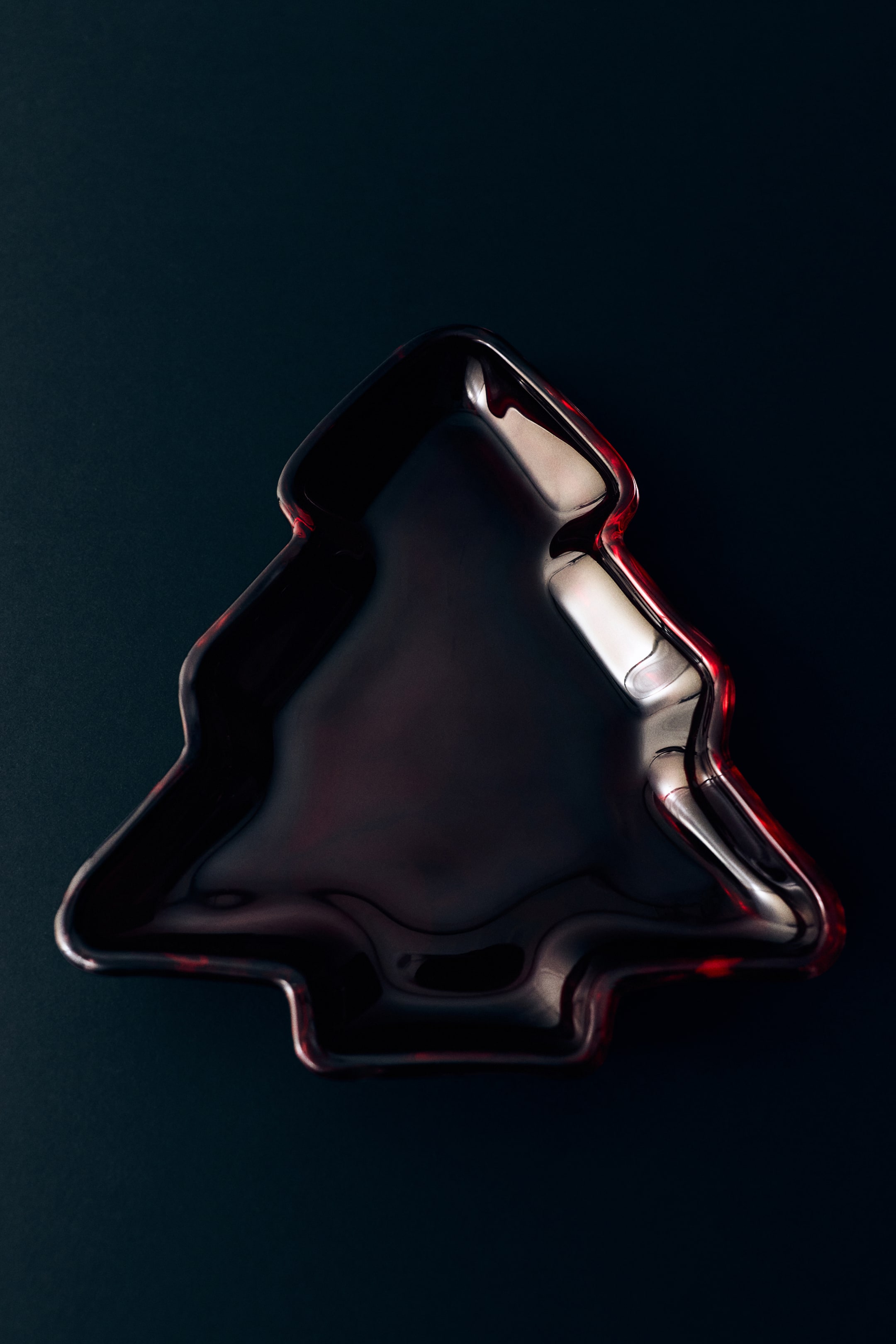 Ver imagen más grande: Una bandeja con forma de pino, elaborada en vidrio rojo oscuro, reposa plana, mostrando su superficie brillante y reflectante desde una perspectiva cenital. La bandeja presenta un borde elevado que define su silueta contorneada, con reflejos rojos que resplandecen a lo largo de los bordes.