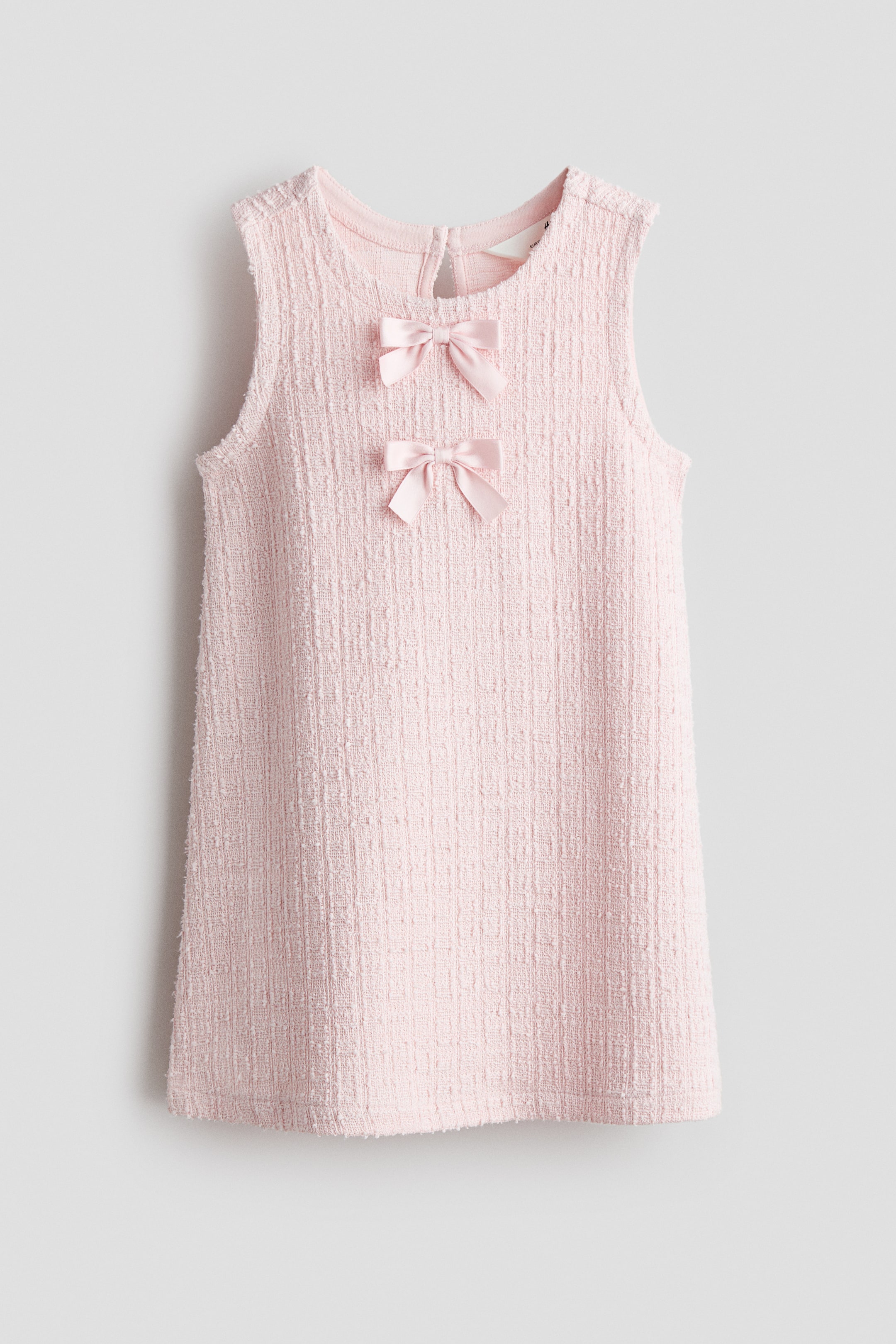 View larger image: A pink sleeveless dress is shown from the front, featuring a textured, tweed-like fabric. Two pink satin bows adorn the upper front bodice, while a round neckline with a keyhole opening and button closure at the back of the neck is visible.