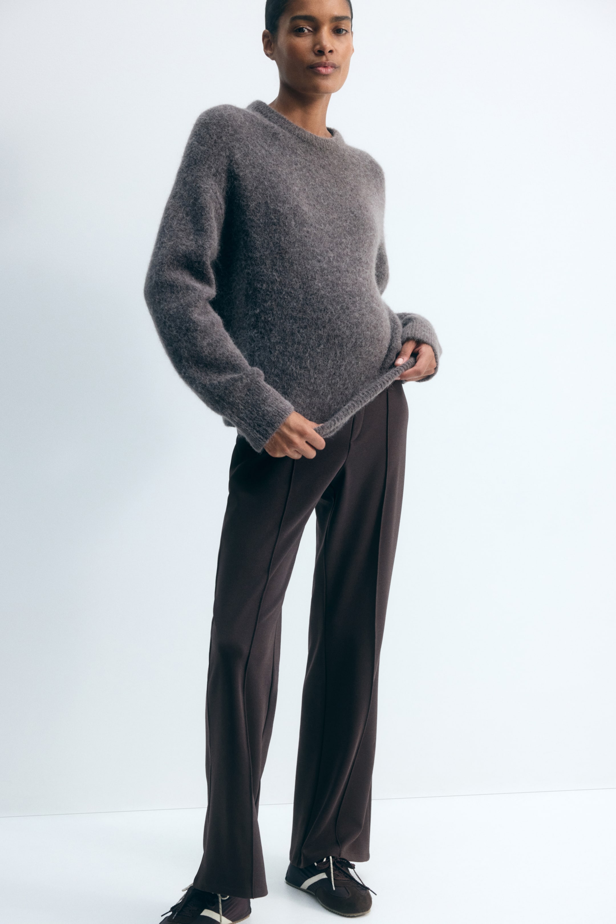 Προβολή μεγαλύτερης εικόνας: Παντελόνι από βαριά καφέ φανέλα, με μπατζάκια bootcut και ραμμένες πιέτες στο μπροστινό μέρος, φορεμένο από το μοντέλο. Το μοντέλο κρατάει το τελείωμα ενός υφασμένου γκρι πουλόβερ, το οποίο φοριέται σε στρώσεις πάνω από τη μέση του παντελονιού.