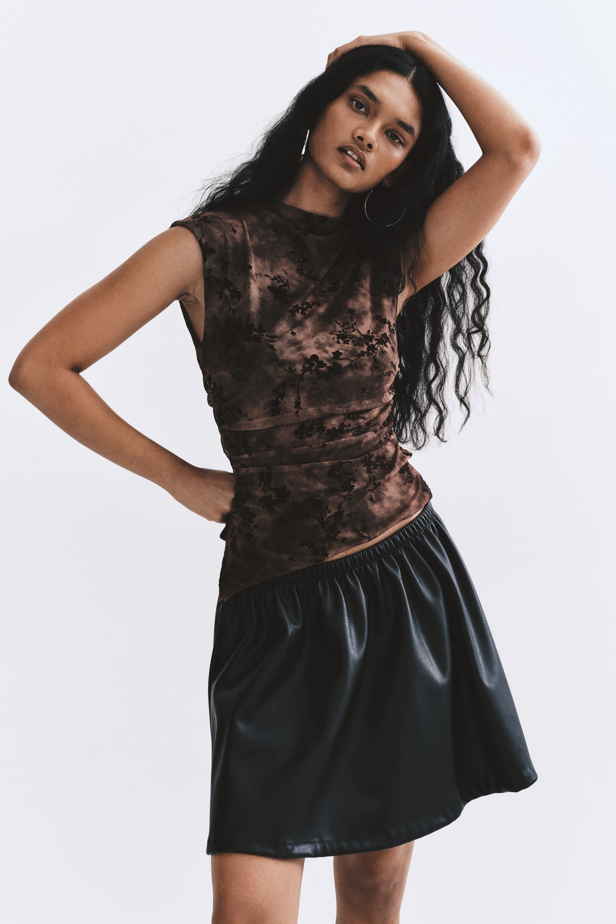 View larger image: A sleeveless, fitted mesh top in a brown tie-dye pattern with a black floral flock print is worn by the model. The top features a high neck, gathered side seams, and an asymmetrical hem, and is styled with a black faux-leather skirt.
