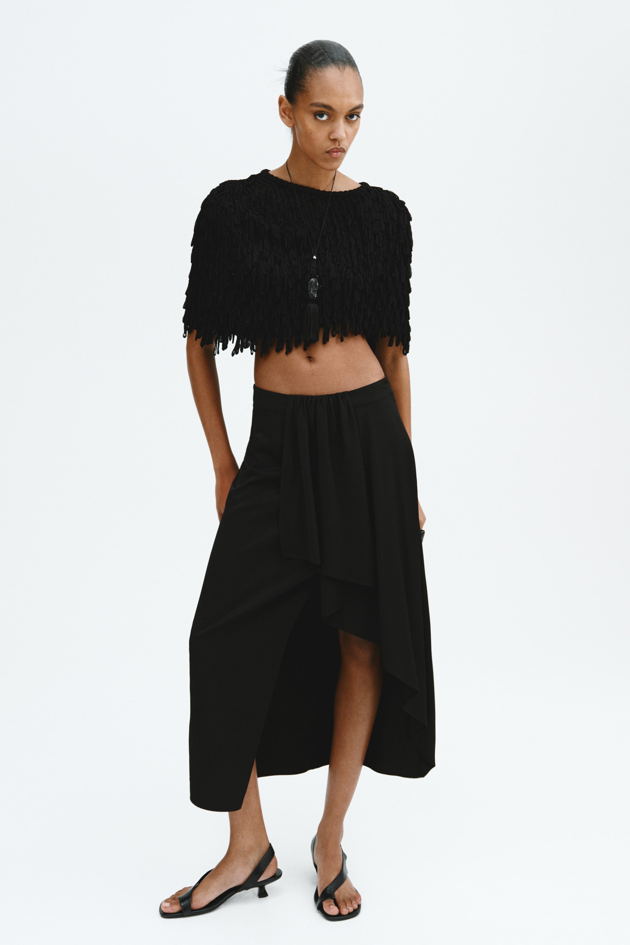 Agrandir l'image: Un crop top noir présente un col rond et est recouvert de franges denses et ébouriffées, tandis qu'une jupe longue noire arbore un ourlet asymétrique haut-bas et un panneau avant drapé et superposé.
