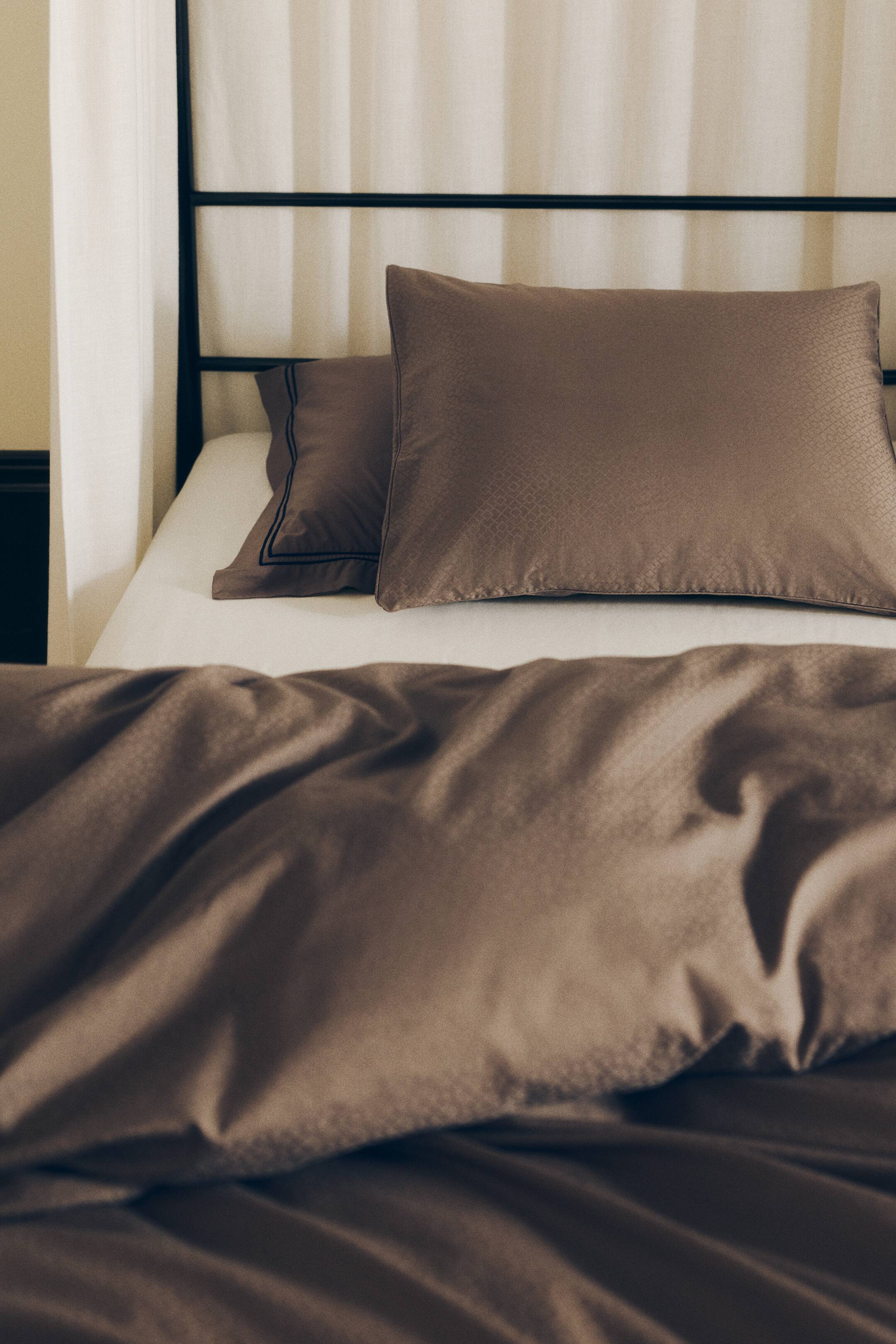 View larger image: A brown cotton sateen duvet cover set is showcasing a matching pillowcase and a draped duvet cover featuring a subtle jacquard-woven geometric pattern. The fabric is displaying a soft sheen that highlights the textured weave of the bedding.