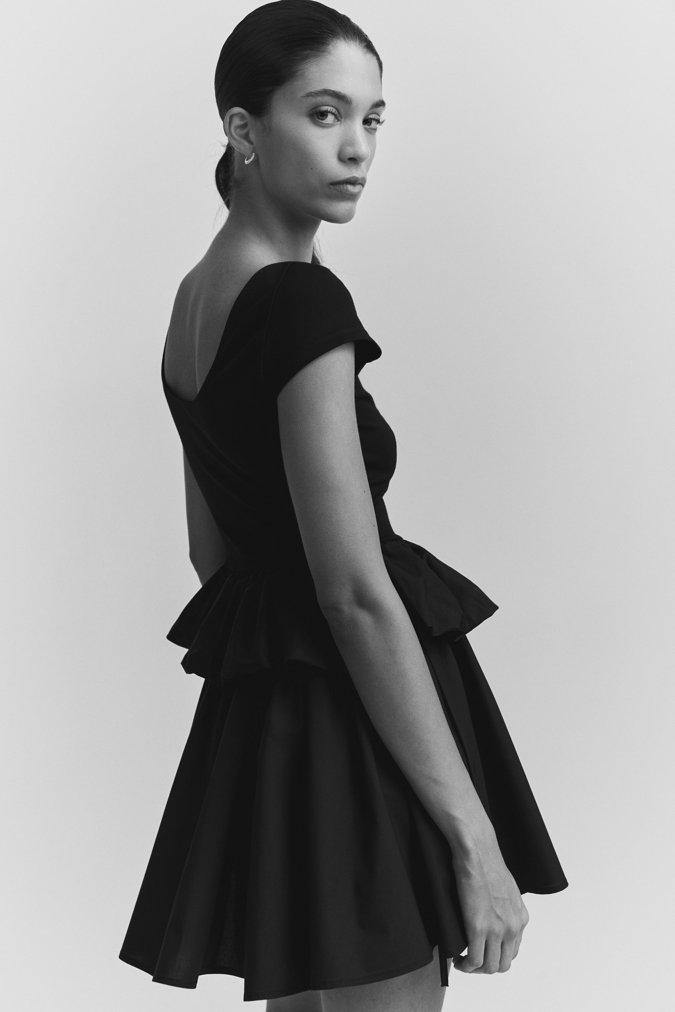 View larger image: A black dress features a wide scoop neckline at the back and short sleeves. It showcases a tiered peplum at the waist that flows into a full, flared short skirt, and is presented from a partial back and side view.