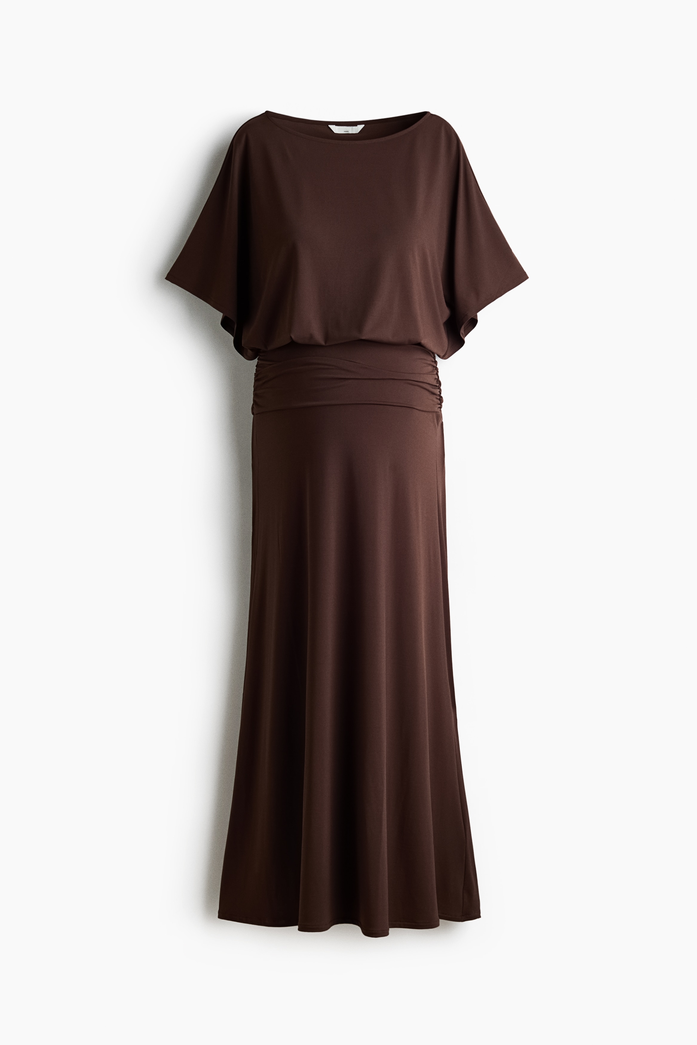 A long, dark brown jersey dress featuring a boat neckline, short sleeves, a loose-fit bodice with a gathered seam, and a fitted, draped section across the tummy is being worn by the model. The model is posing with one knee raised, showcasing the drape and movement of the gently flared skirt.