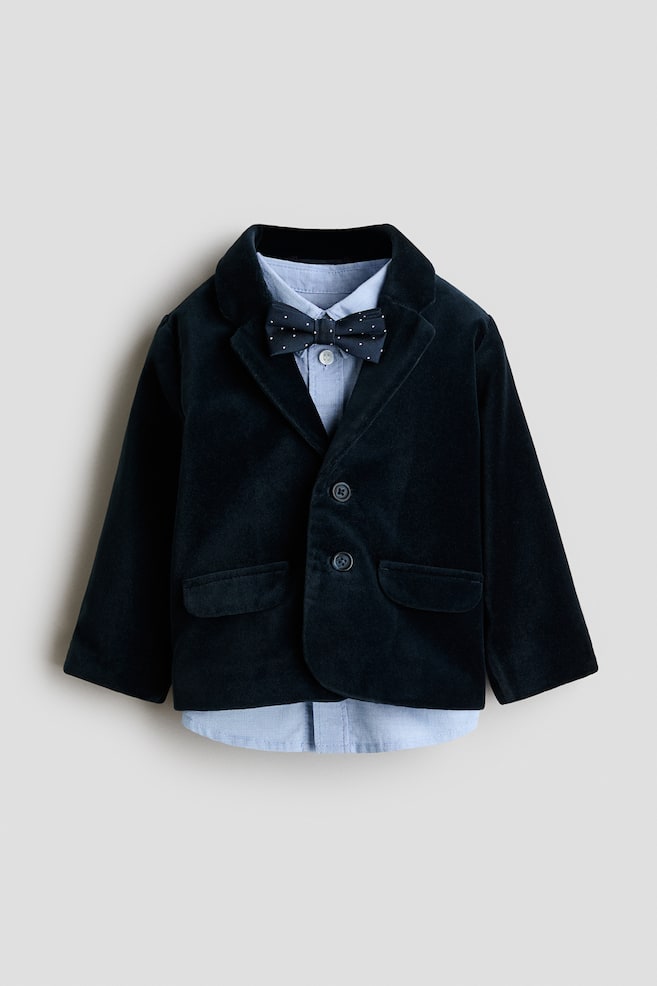 Et barns formelle outfit vises forfra og bestÃ¥r af en mÃ¸rk marineblÃ¥ blazer i flÃ¸jl, bÃ¥ret over en lyseblÃ¥ skjortebluse med knapper og en matchende marineblÃ¥ butterfly med prikker. Blazeren har indhakrevers, lukning med to knapper foran og to lommer med klap, mens skjortens kant stikker en smule frem under den nederste kant. - 1