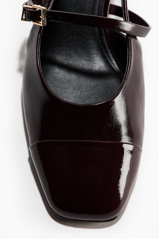 A dark burgundy patent leather Mary Jane shoe is shown in a close-up, front-facing view, highlighting its glossy, high-shine finish. It features a structured square cap toe and a slim, adjustable instep strap with a small, gold-tone rectangular buckle. - 1