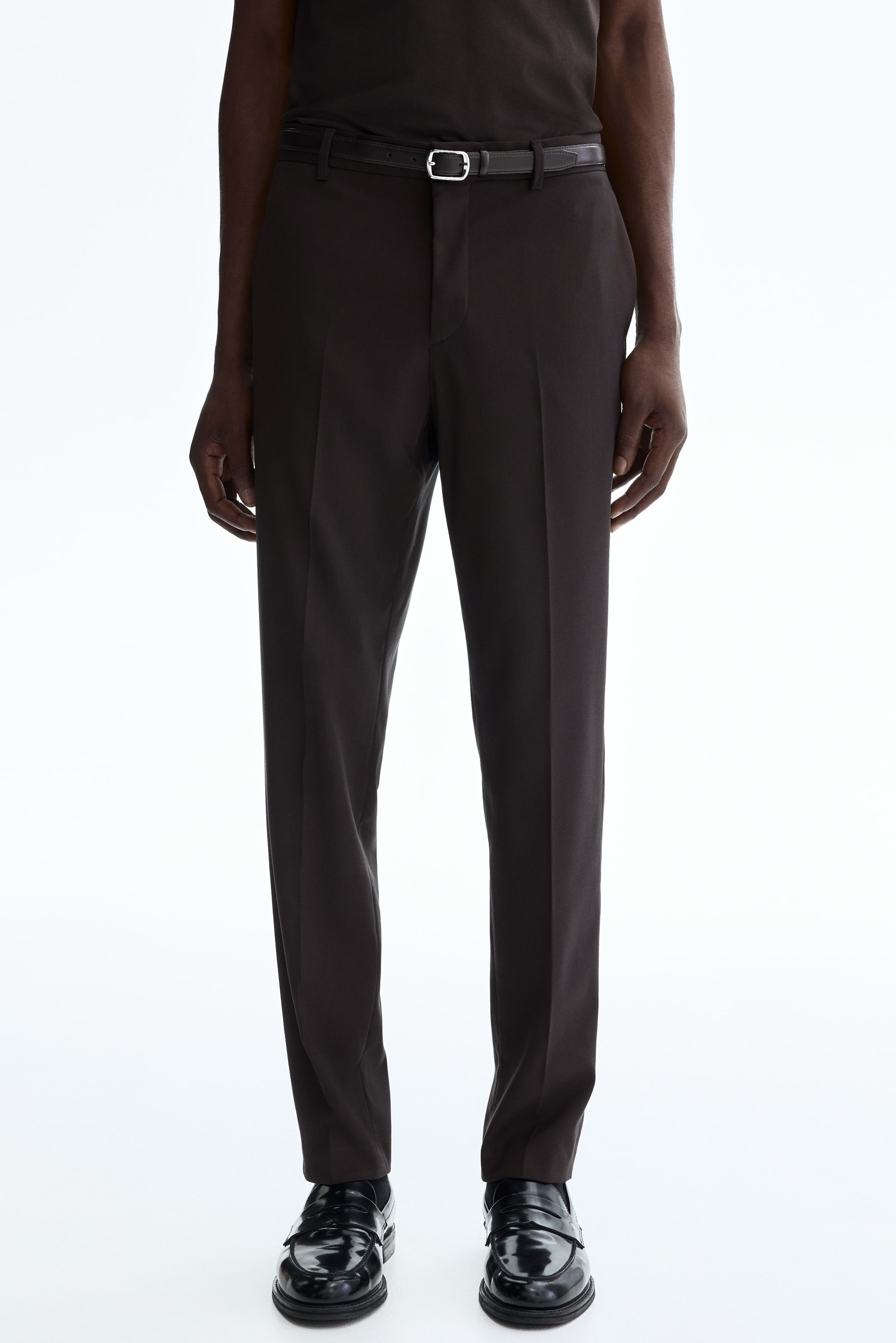 Ampliar la imagen: El modelo lleva pantalones de traje ajustados en color marrón oscuro con pliegues prominentes en las piernas. Un cinturón negro con hebilla plateada se lleva a través de las trabillas del cinturón de los pantalones.