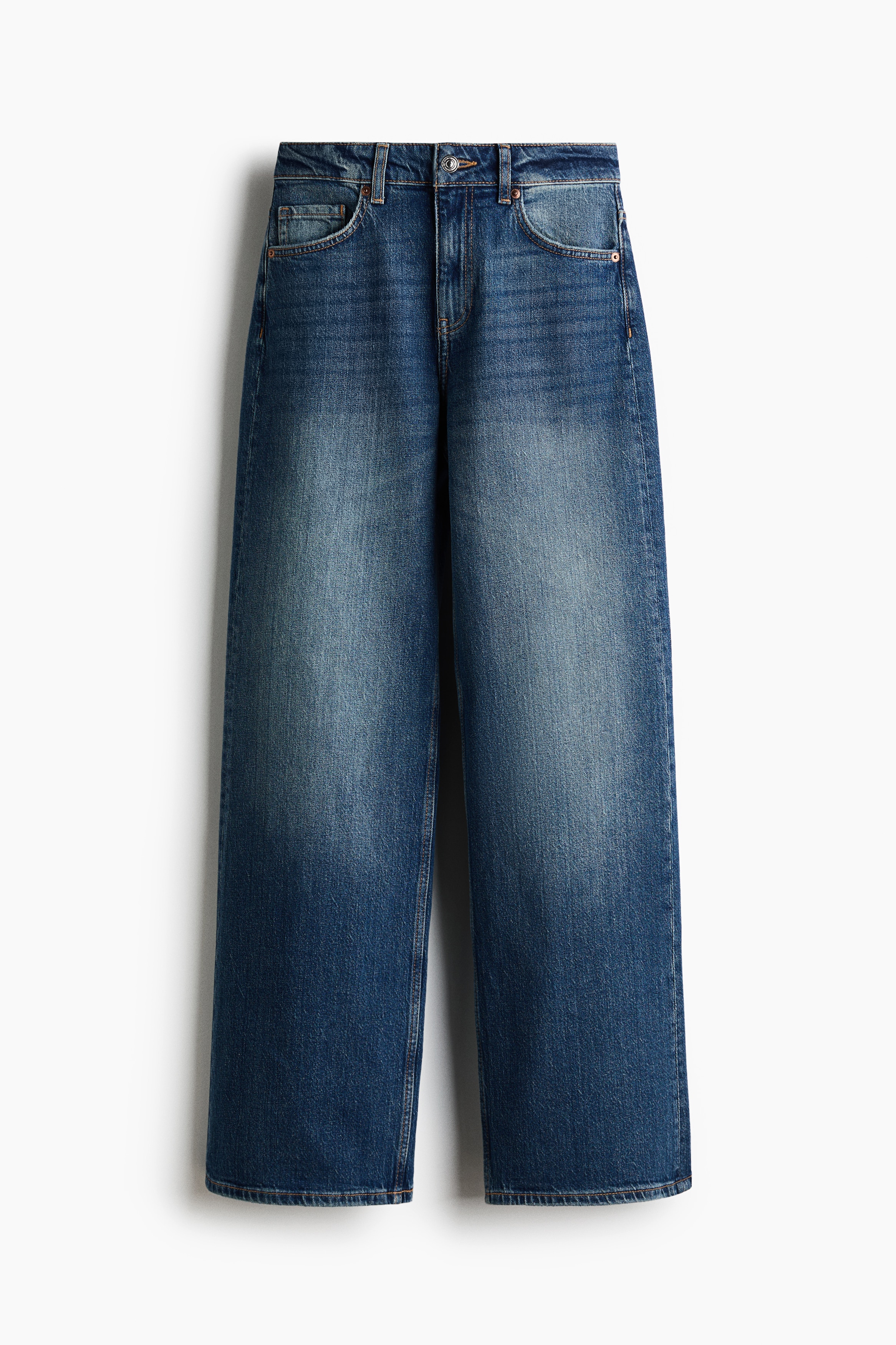 The model is seated on a chair wearing blue denim jeans with a loose, straight-leg fit. The medium-wash jeans are styled with a dark short-sleeved T-shirt and low-top sneakers.