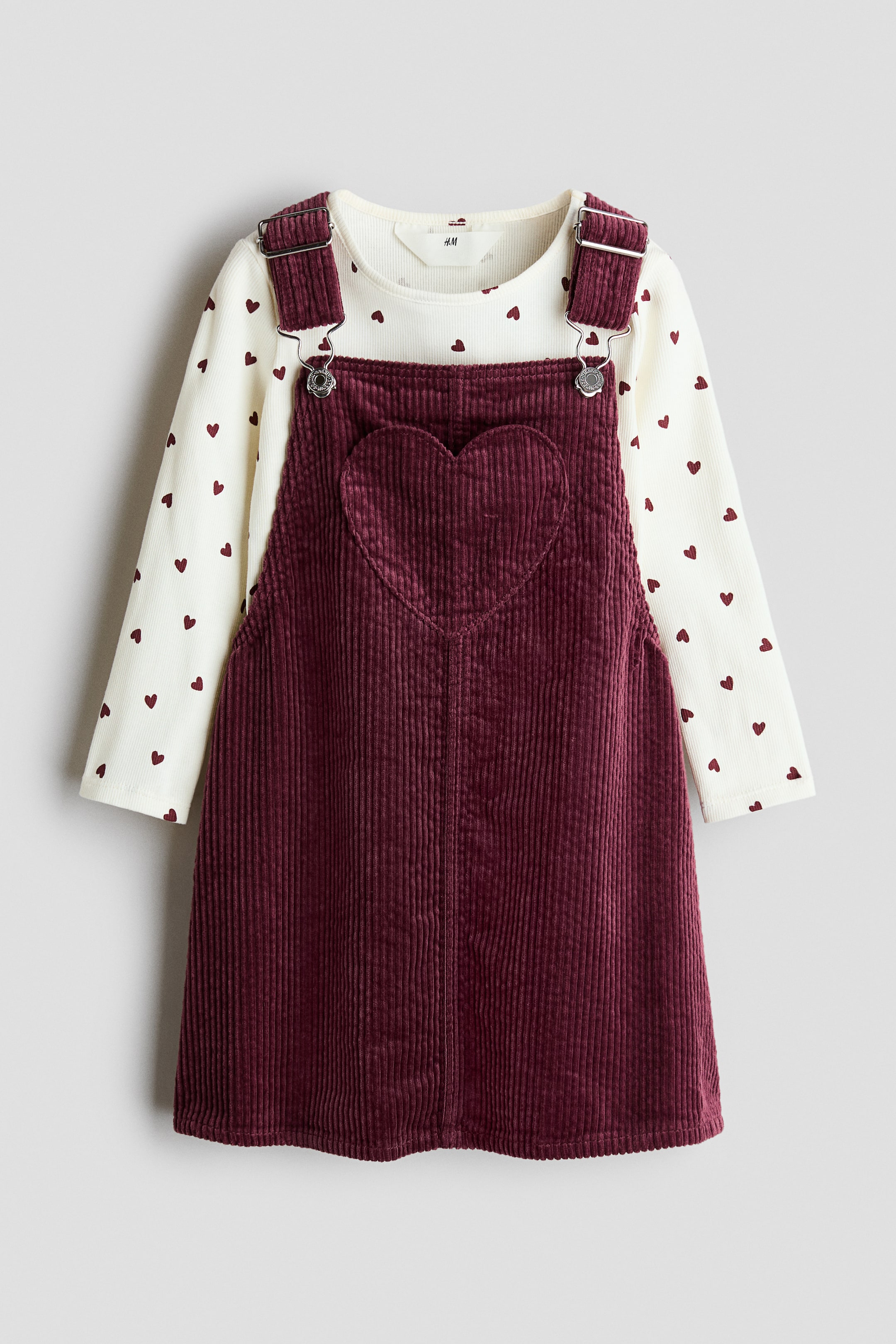 View larger image: A front-facing view of a children's clothing set shows a dark red corduroy dungaree dress layered over a cream-coloured, long-sleeved ribbed top. The dress features adjustable shoulder straps with silver-coloured metal clasps and a heart-shaped chest pocket, while the top displays an all-over pattern of small red hearts.