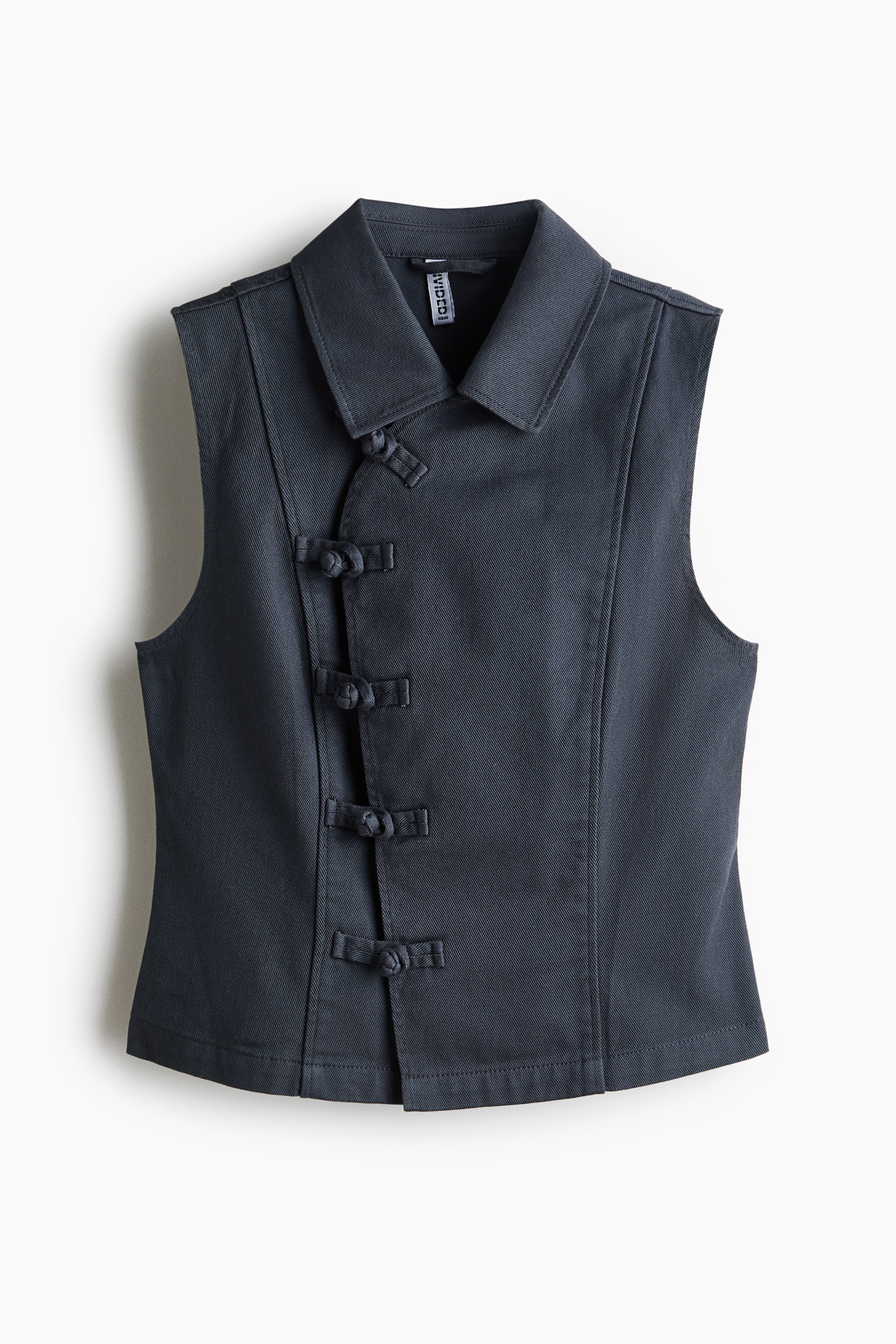 Ampliar la imagen: Se muestra una blusa sin mangas de color gris oscuro vista de frente, con cuello en punta y cinco pares de cierres decorativos tipo frog a lo largo del centro. La prenda presenta una silueta ajustada con detalles de costuras verticales en los paneles delanteros.
