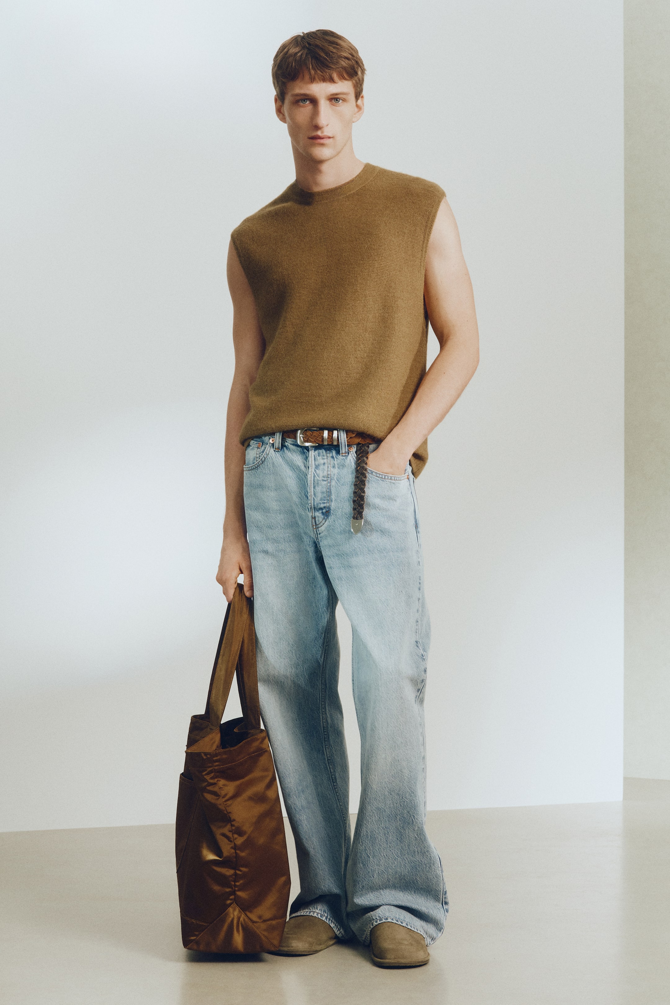 Ampliar la imagen: Un top tejido sin mangas de color marrón se lleva con jeans de corte relajado y lavado claro, asegurados con un cinturón trenzado marrón. Unos zapatos de gamuza marrón completan el atuendo, y un bolso tote marrón brillante se lleva al lado.