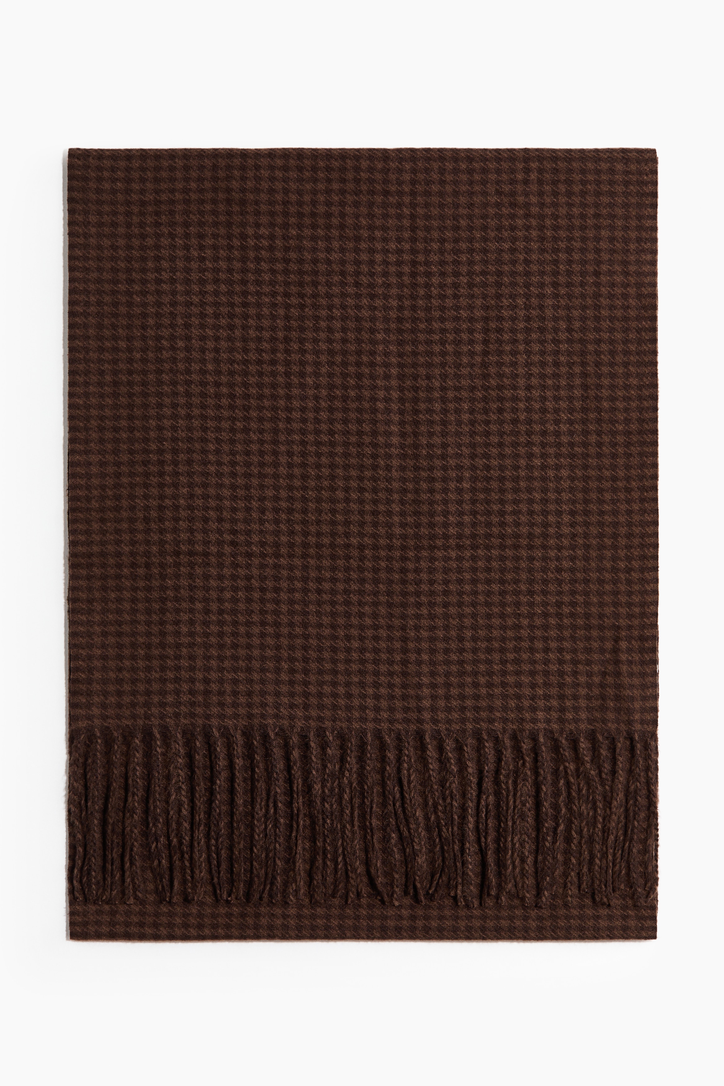 En brun scarf med hundtandsmÃ¶nster i mjuk vÃ¤v bÃ¤rs av modellen, virad runt halsen och neddragen framtill Ã¶ver en mÃ¶rkbrun kappa. Scarfen har tvinnade fransar lÃ¤ngs de korta sidorna.