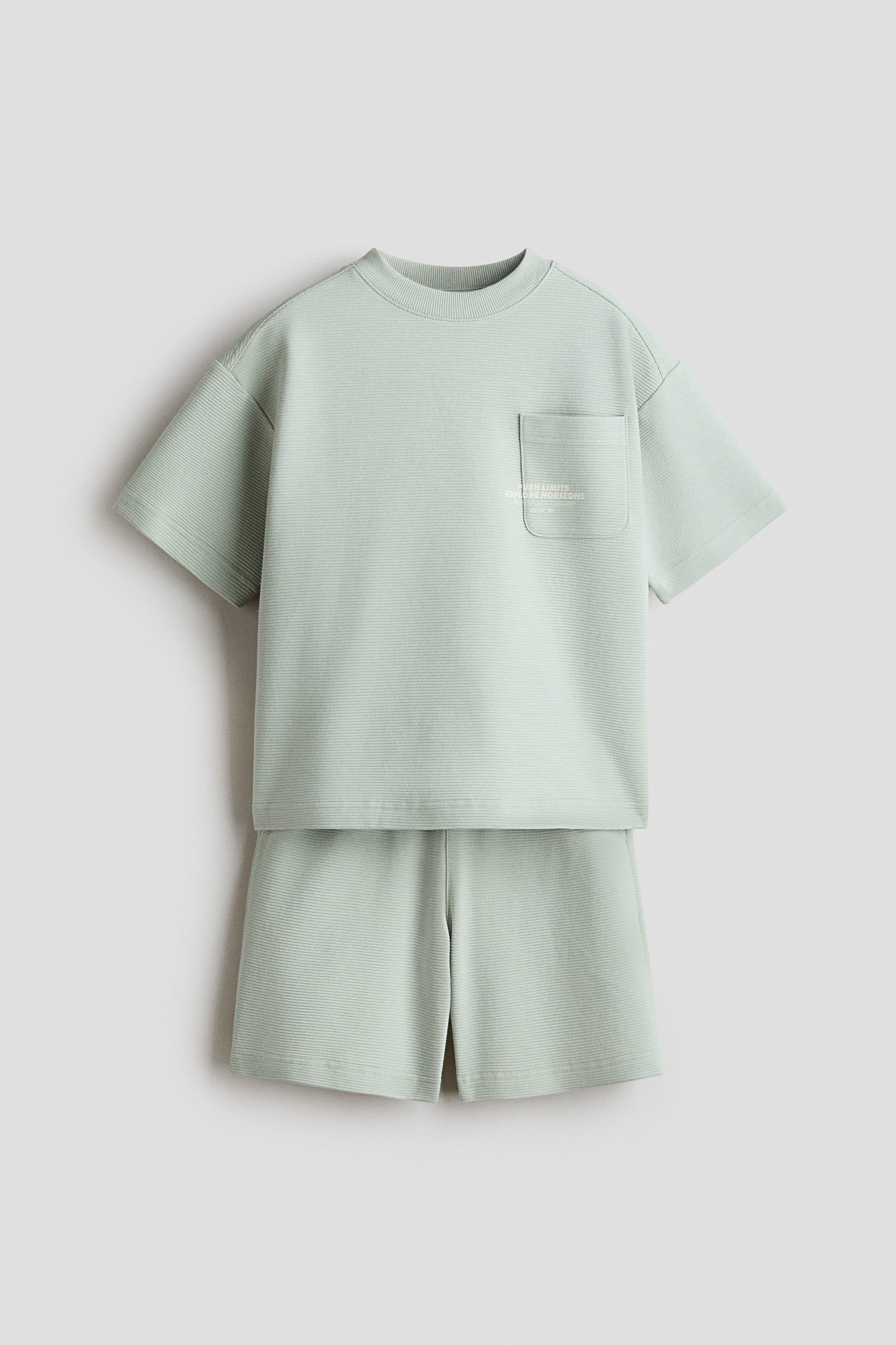 View larger image: A light green short-sleeved T-shirt dan set seluar pendek yang sepadan dipamerkan dari depan. Kedua-dua bahagian menampilkan tekstur berusur halus, dengan T-shirt termasuk leher bulat dan poket tampal di dada kiri yang menunjukkan teks samar.