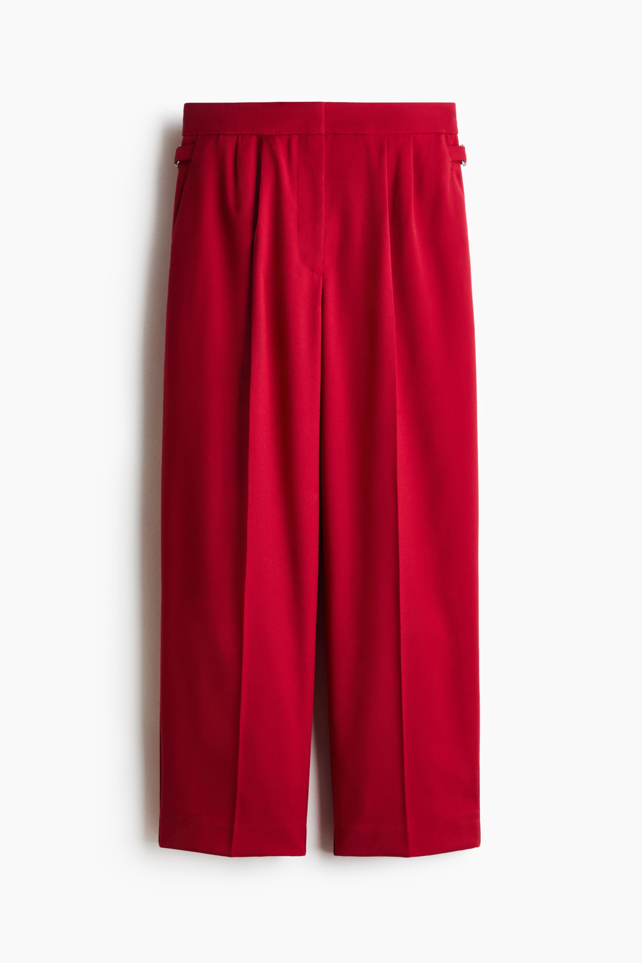 View larger image: A front view of red woven trousers shows a high-waisted design with straight legs, featuring front pleats and sharp creases down the centre. The trousers display a flat-front waistband with a concealed closure and small adjustable tabs with metal buckles on the sides.