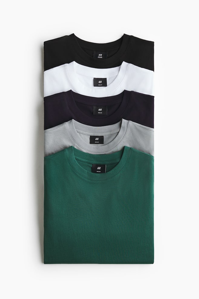 Vijf opgevouwen katoenen jersey T-shirts liggen in een verticale stapel, van voren gezien, waarbij hun ronde halslijnen met ribboord zichtbaar zijn in zwart, wit, donkerpaars, grijs en bosgroen. - 1