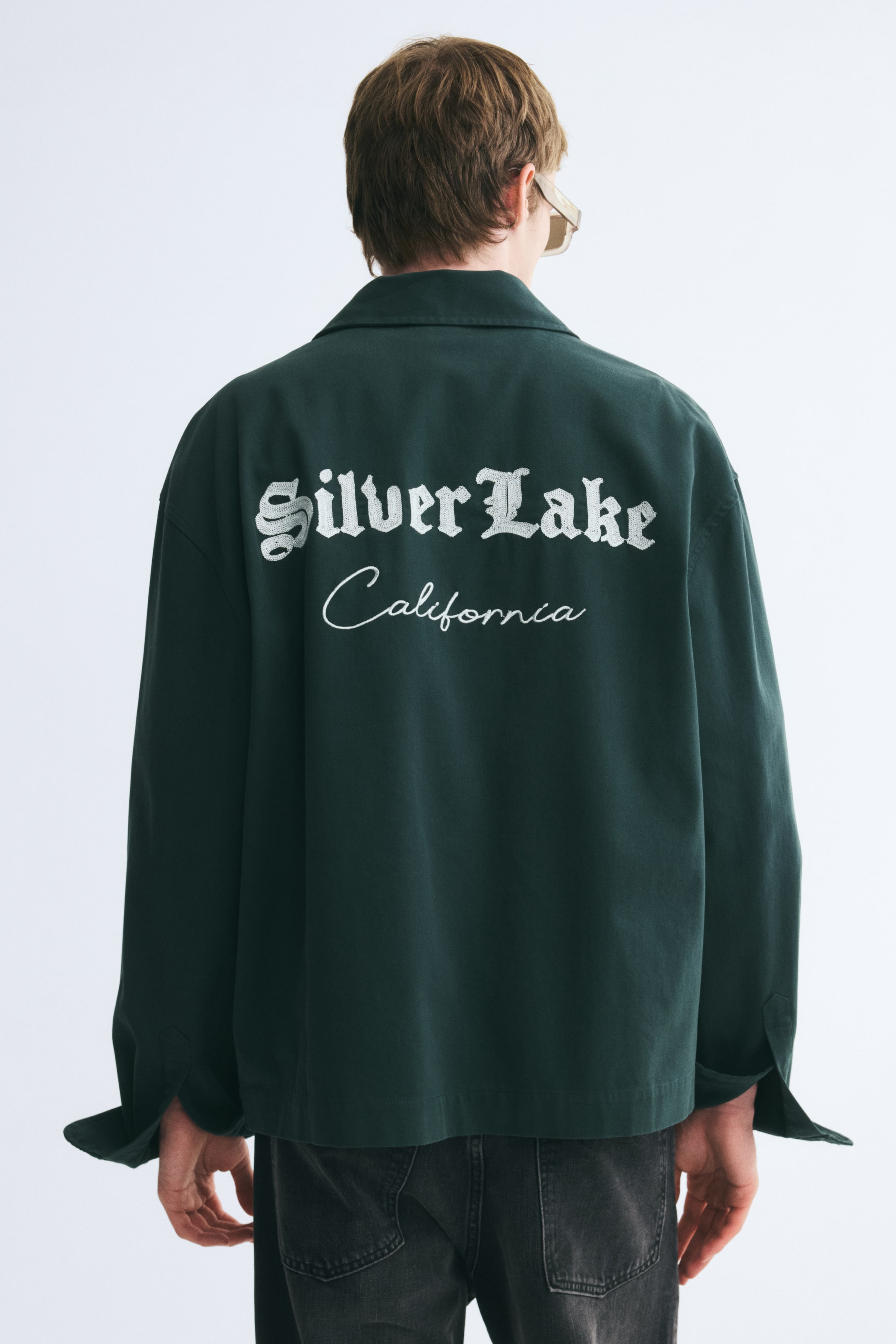 View larger image: The model is wearing a dark green cotton twill overshirt, shown from the back. The overshirt features white text on the back reading "Silver Lake" in a gothic font and "California" in cursive script, and is paired with dark grey jeans.