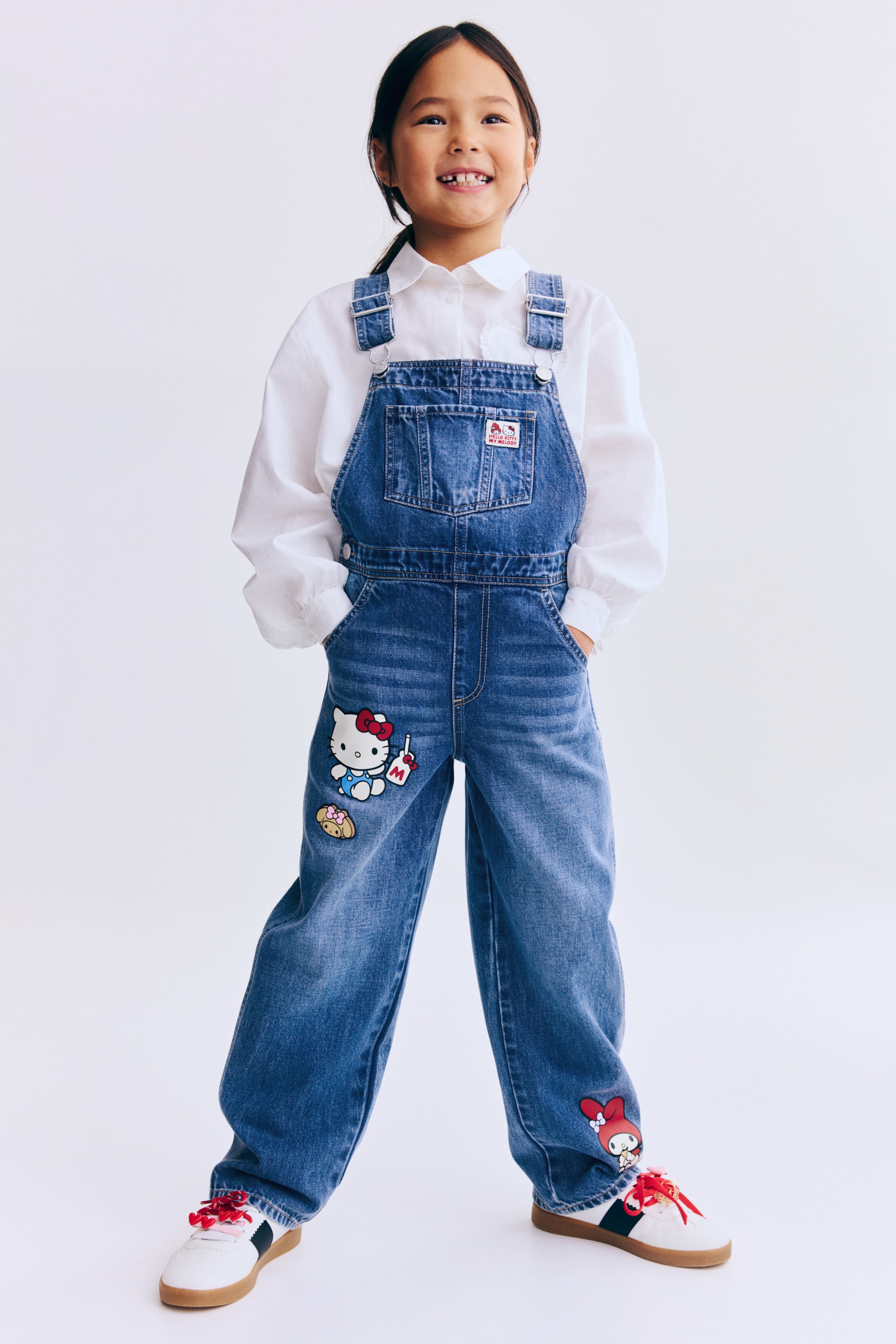 Se større billede: Blå denimoveralls har et bryststykke foran med en "Hello Kitty with My Melody"-patch på lommen, justerbare skulderstropper og knapper i siderne. Store Hello Kitty- og andre karakterpatches pryder de vide ben, som sidder løst over en hvid, langærmet skjorte med krave.