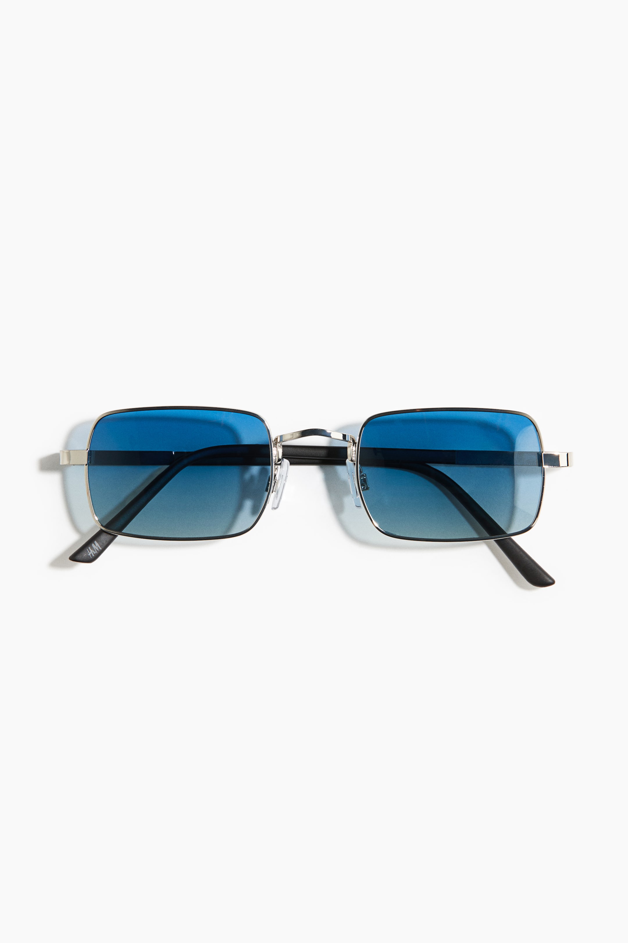 Agrandir l'image: Les lunettes de soleil sont présentées en vue de face, avec une monture rectangulaire en métal de ton argenté. Elles arborent des verres dégradés teintés de bleu, passant d'un bleu plus foncé en haut à une teinte plus claire en bas, avec des branches noires s'étendant sur les côtés.