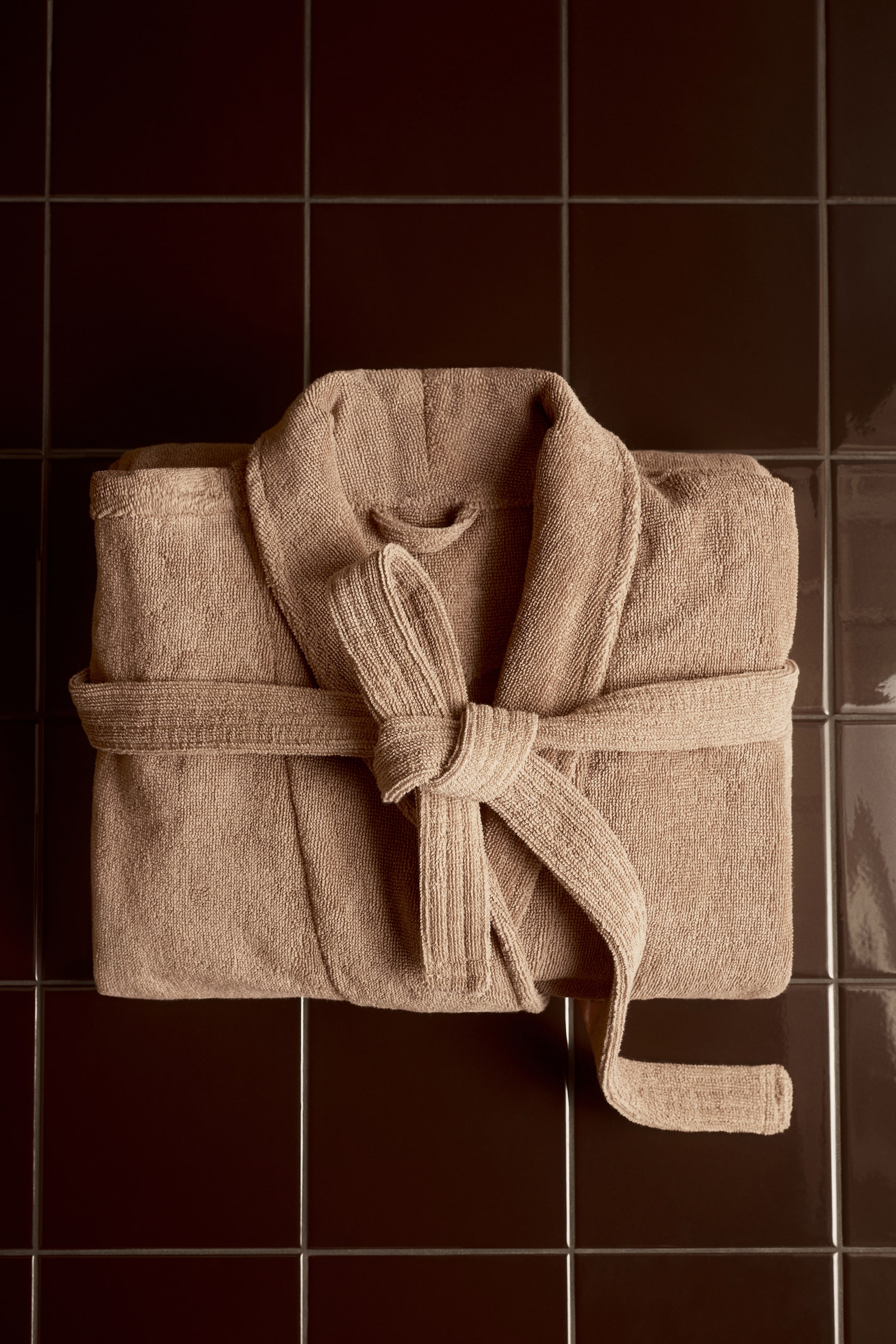 View larger image: A tan terry cloth bathrobe lies flat, neatly folded, with its belt tied in a bow at the front.
