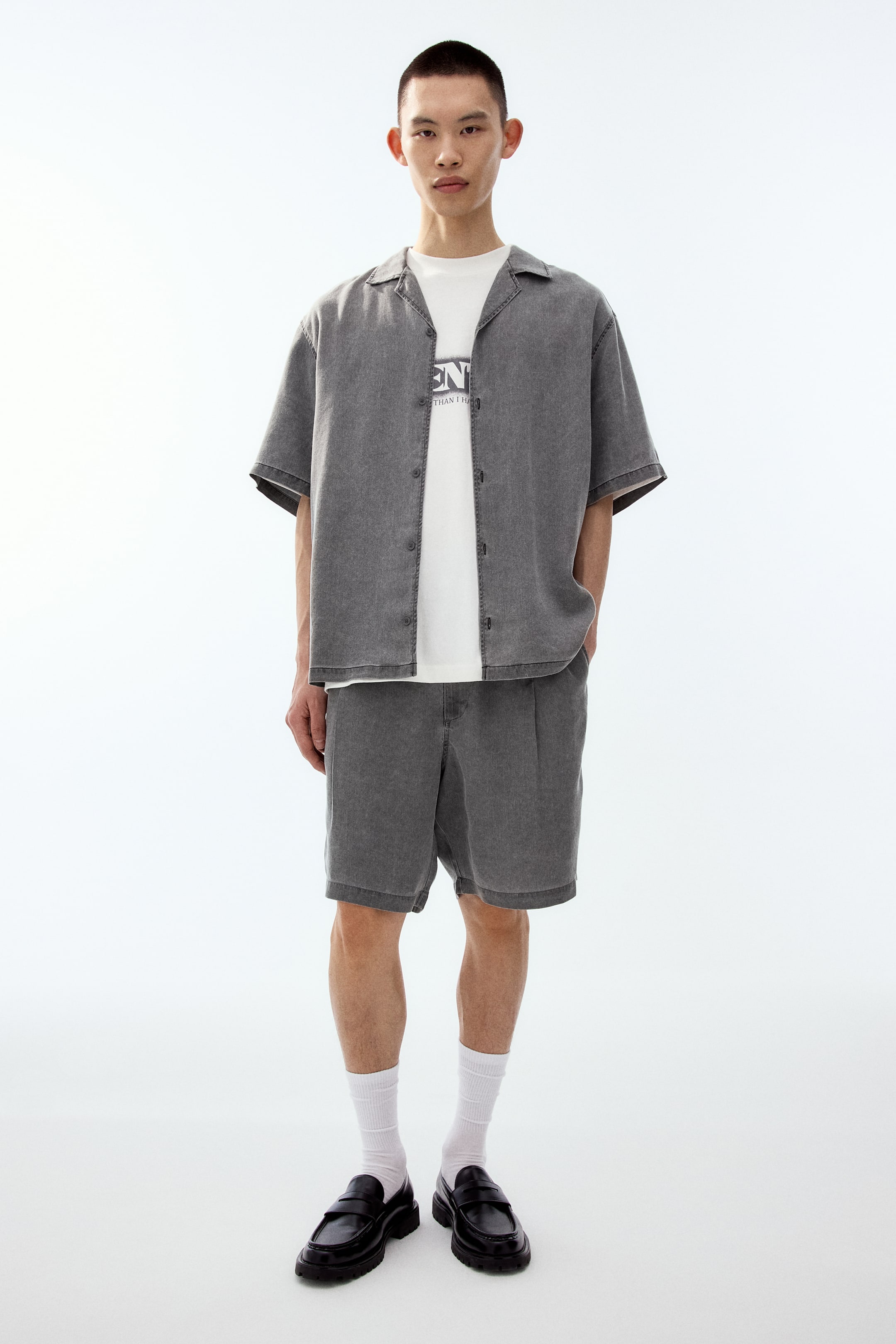 Xem ảnh lớn hơn: Chiếc áo sơ mi tay ngắn màu xám acid-washed, có cổ revere và cúc phía trước, được mặc hờ khoác ngoài áo thun trắng. Quần short xếp ly cùng tông xám acid-washed có dáng relaxed fit, rộng thoải mái và có độ dài trên đầu gối.