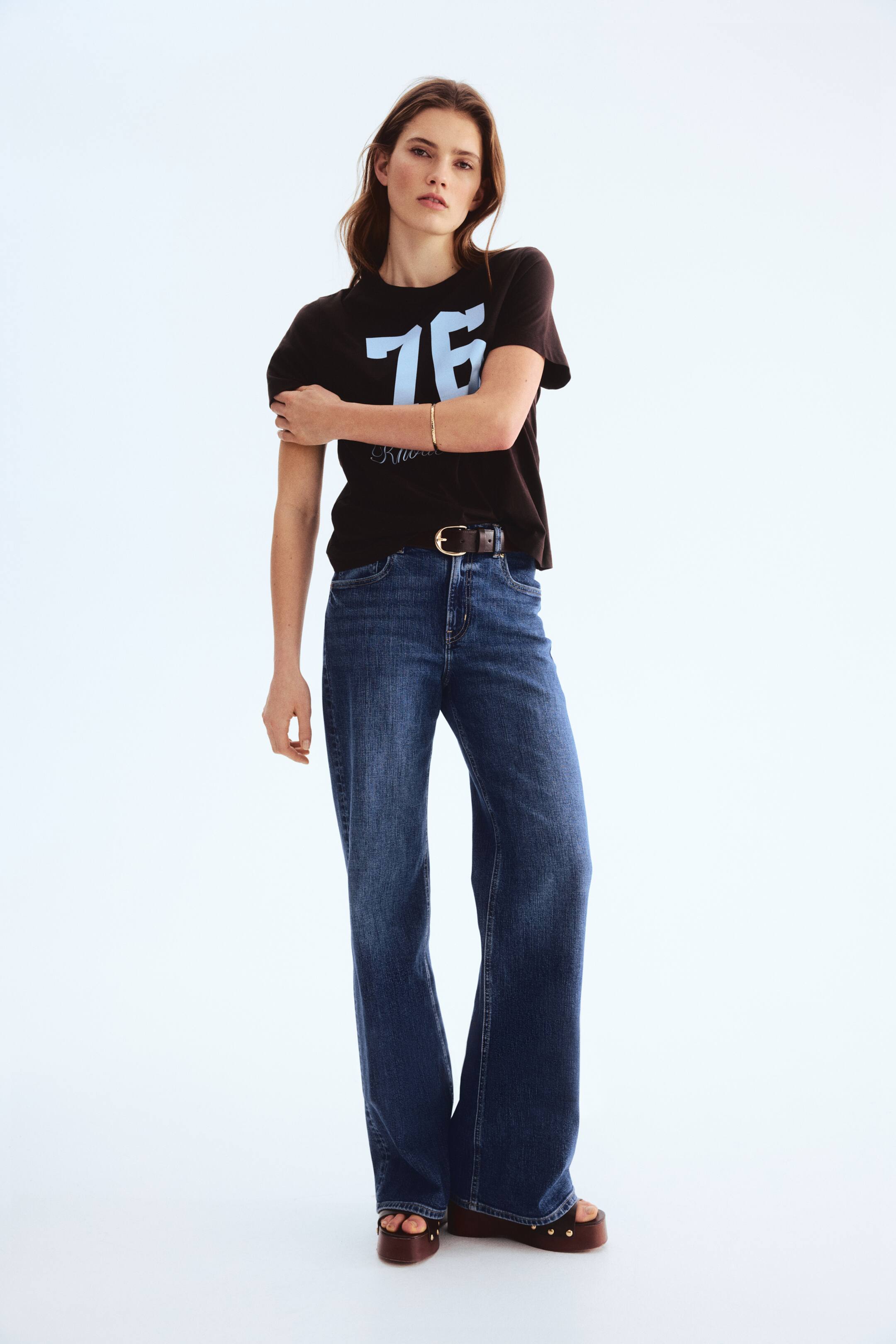 View larger image: A dark-hued T-shirt showcasing a light blue "76" print on the front, complemented by a round, rib-trimmed neckline, is being worn. The model is gently holding her left sleeve, and the T-shirt is partially tucked into blue jeans.