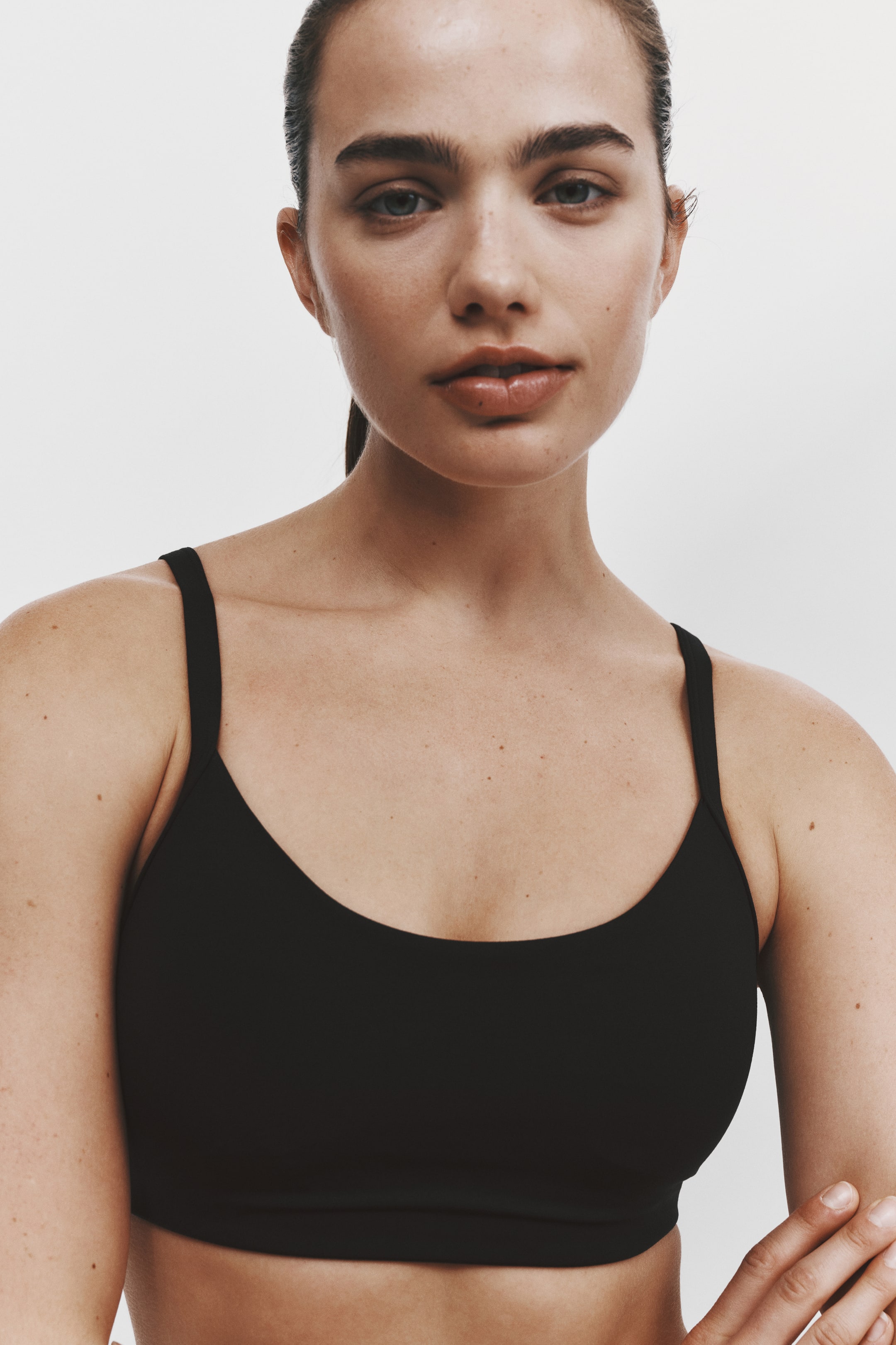 Visualizza immagine più grande: Un reggiseno sportivo nero con scollo rotondo e sottili spalline è indossato da una modella in un primo piano frontale. Il capo presenta una texture liscia e senza cuciture e un’ampia fascia sottoseno di supporto.