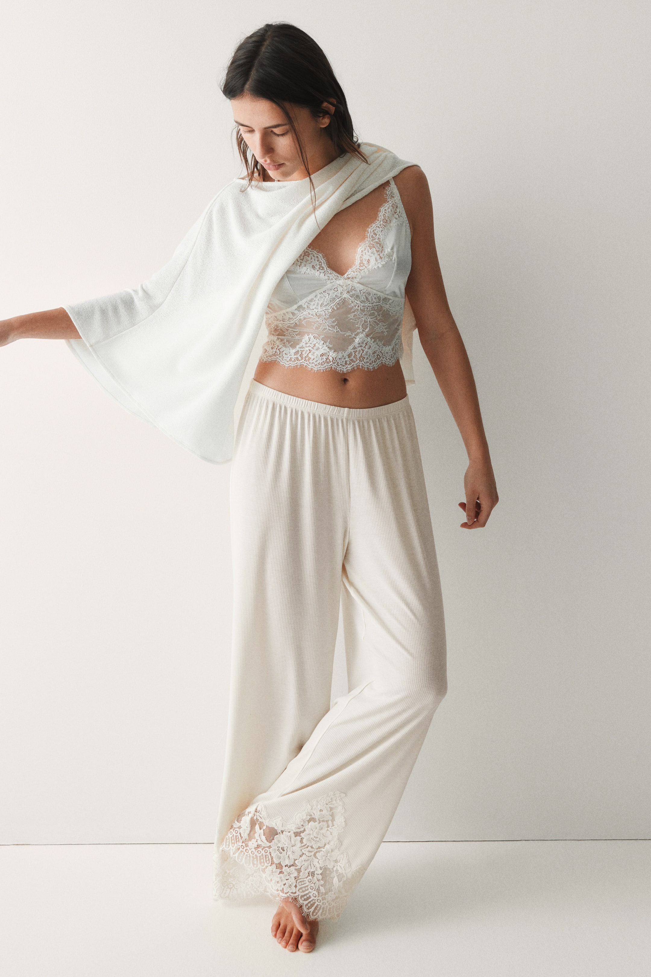 Voir une image plus grande: Un ensemble de détente couleur crème est présenté, comprenant un bralette en dentelle avec des bonnets triangles et des bords festonnés, un pantalon à jambes larges en maille côtelée avec des inserts en dentelle aux poignets extérieurs, et un cache-cœur assorti en maille côtelée.