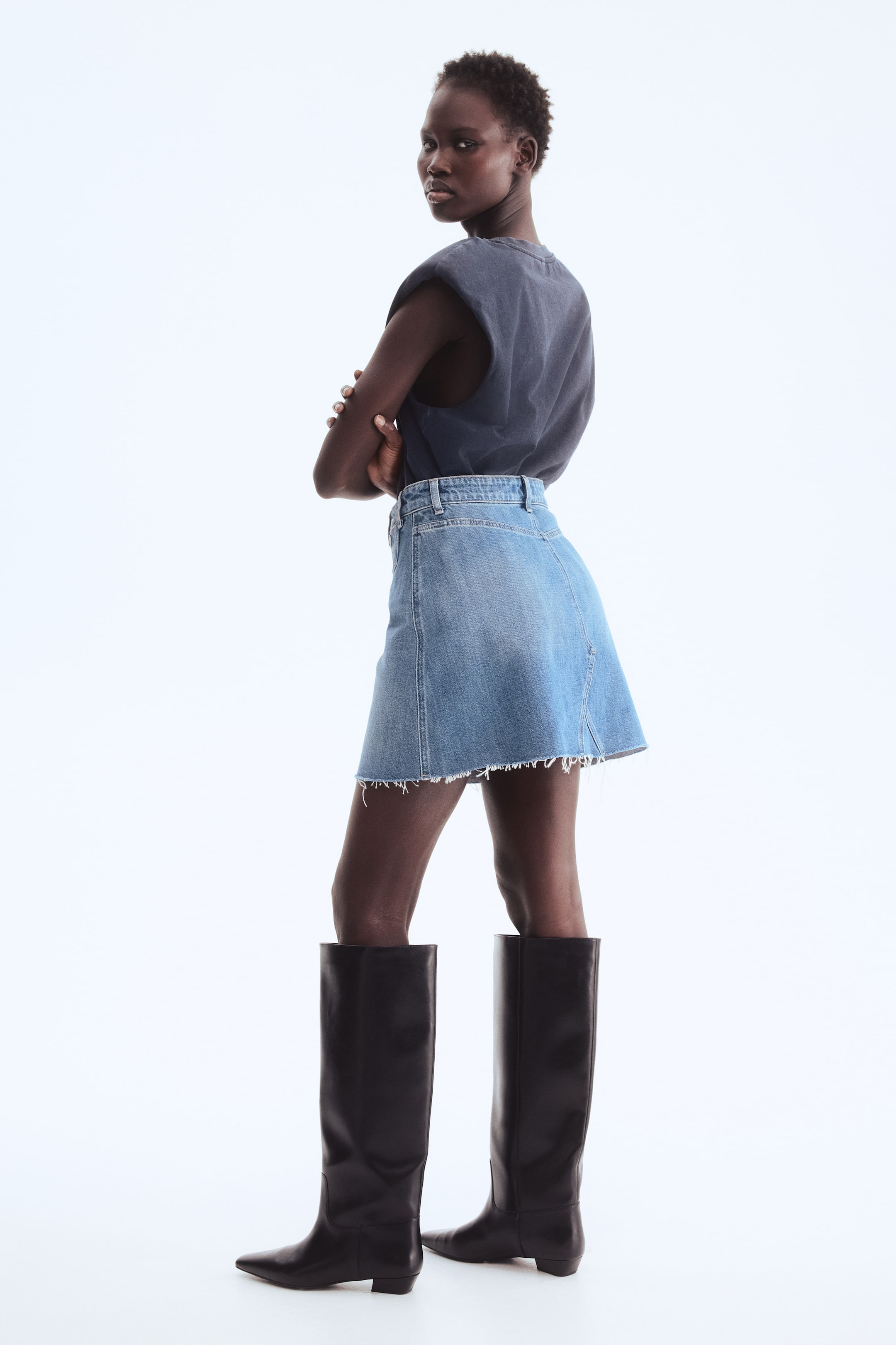 View larger image: A light blue denim A-line skirt is being showcased from a slightly rear-side angle, highlighting a raw, frayed hem, visible belt loops at the waist, and a single back pocket.