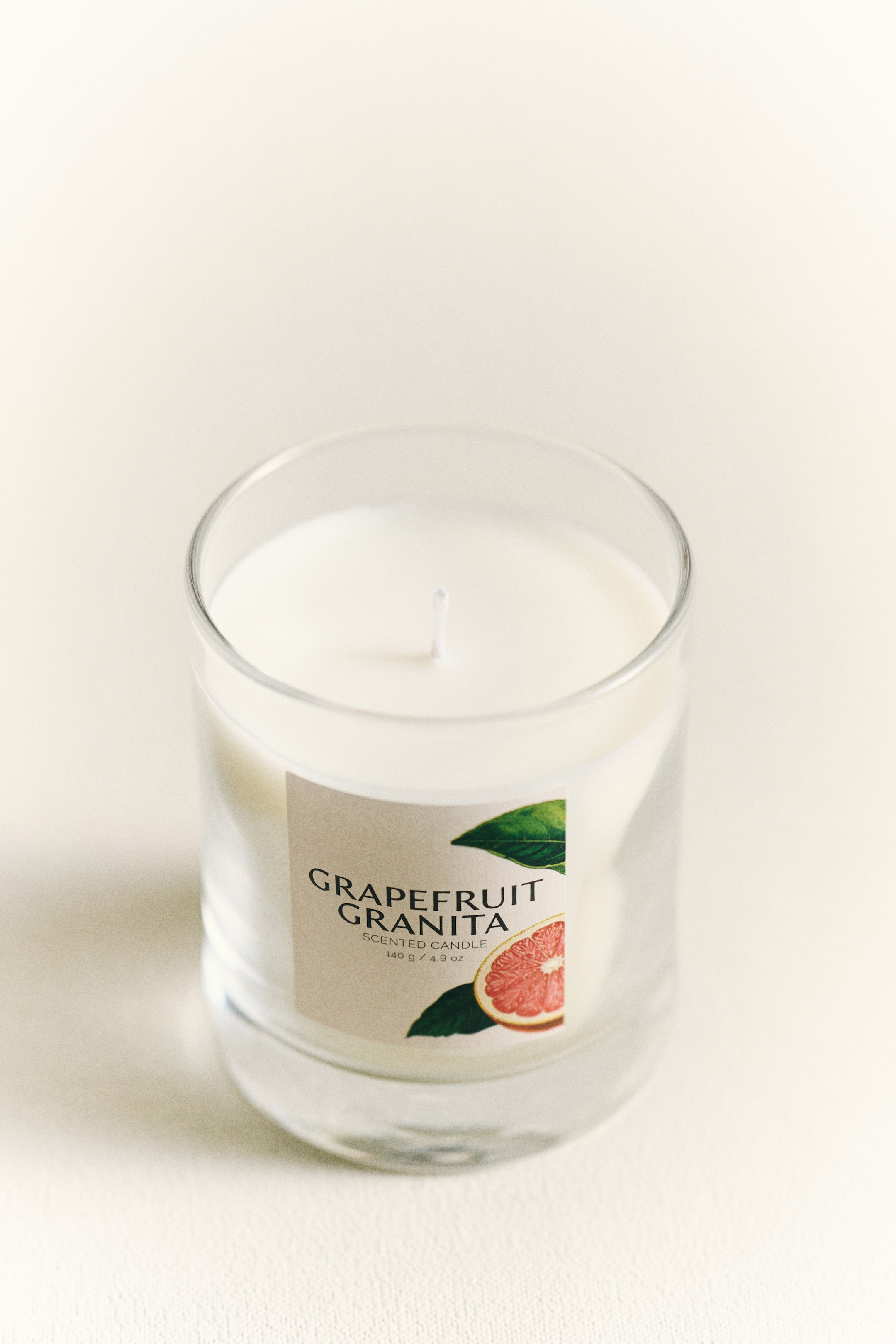 View larger image: A scented candle is showcased in a clear glass jar, angled to reveal its top and front, with white wax filling the jar and a single white wick rising from the centre. A light-coloured label on the front displays "GRAPEFRUIT GRANITA", "SCENTED CANDLE", "140 g / 4.9 oz", and a grapefruit illustration.