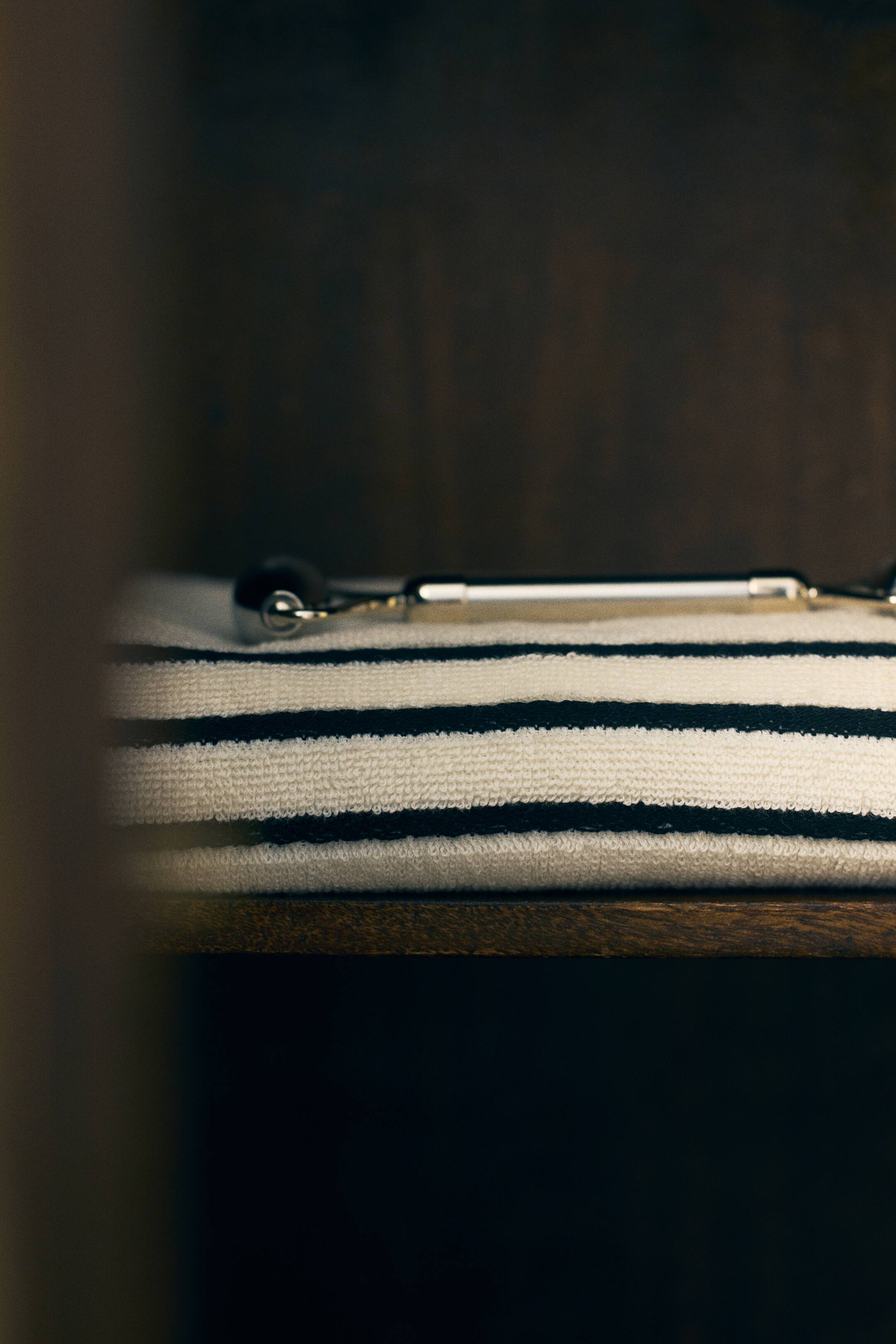 Agrandir l'image: Un torchon plié en éponge de coton blanc doux, orné de rayures noires formant un motif quadrillé minimaliste, repose sur une étagère en bois. Un rouleau de massage métallique est posé sur le torchon plié.