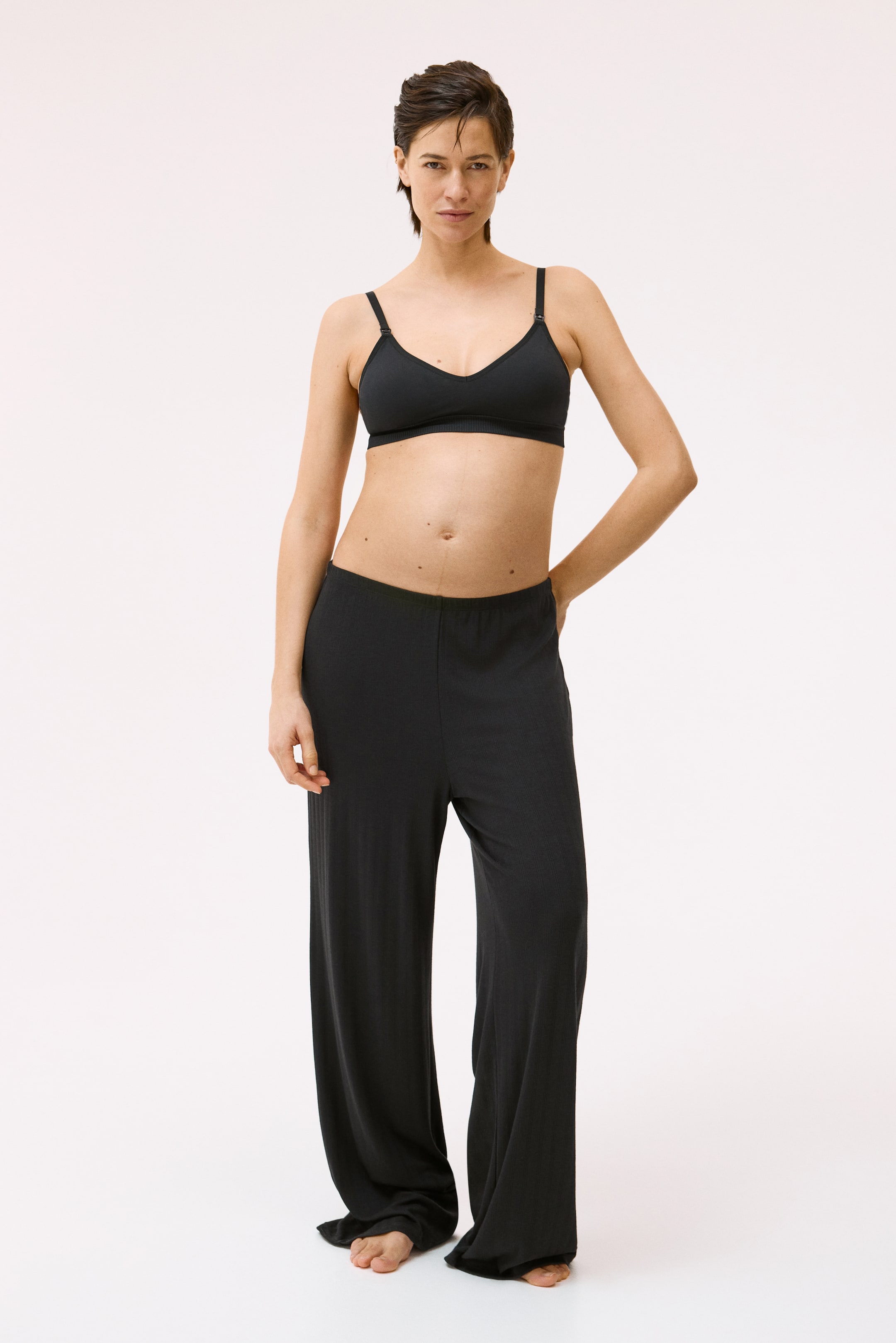 View larger image: A black bra featuring smooth cups, a V-neckline, thin adjustable shoulder straps, and a wide ribbed underband. It is paired with coordinating black wide-leg trousers, showcasing a high waist and a subtle vertical texture.