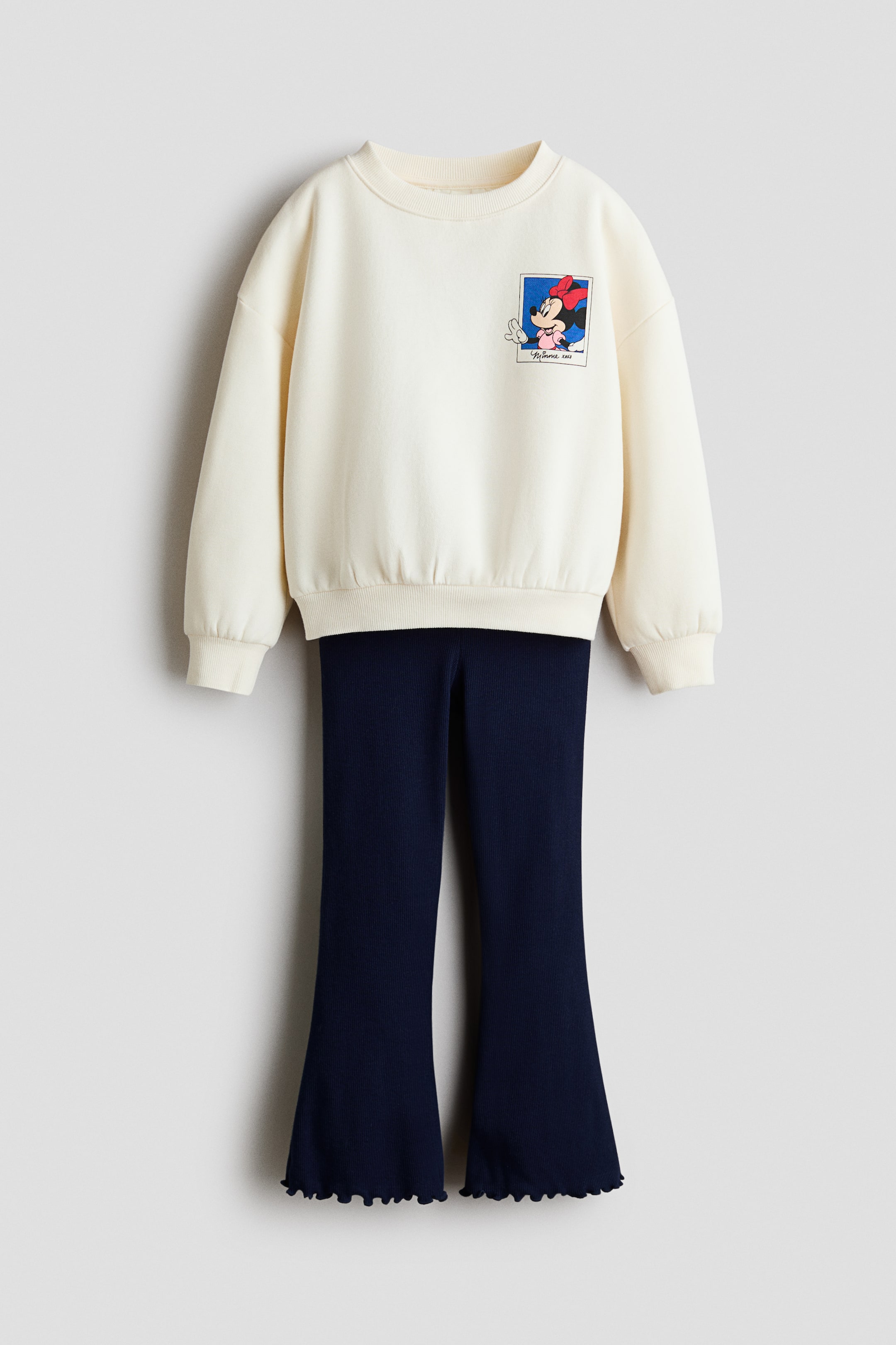 View larger image: A cream crew neck sweatshirt features a rectangular graphic print of Minnie Mouse in a red bow, waving, on the left chest, with ribbed cuffs and hem. It is layered over dark blue ribbed flare pants showcasing a ruffled lettuce edge hem.