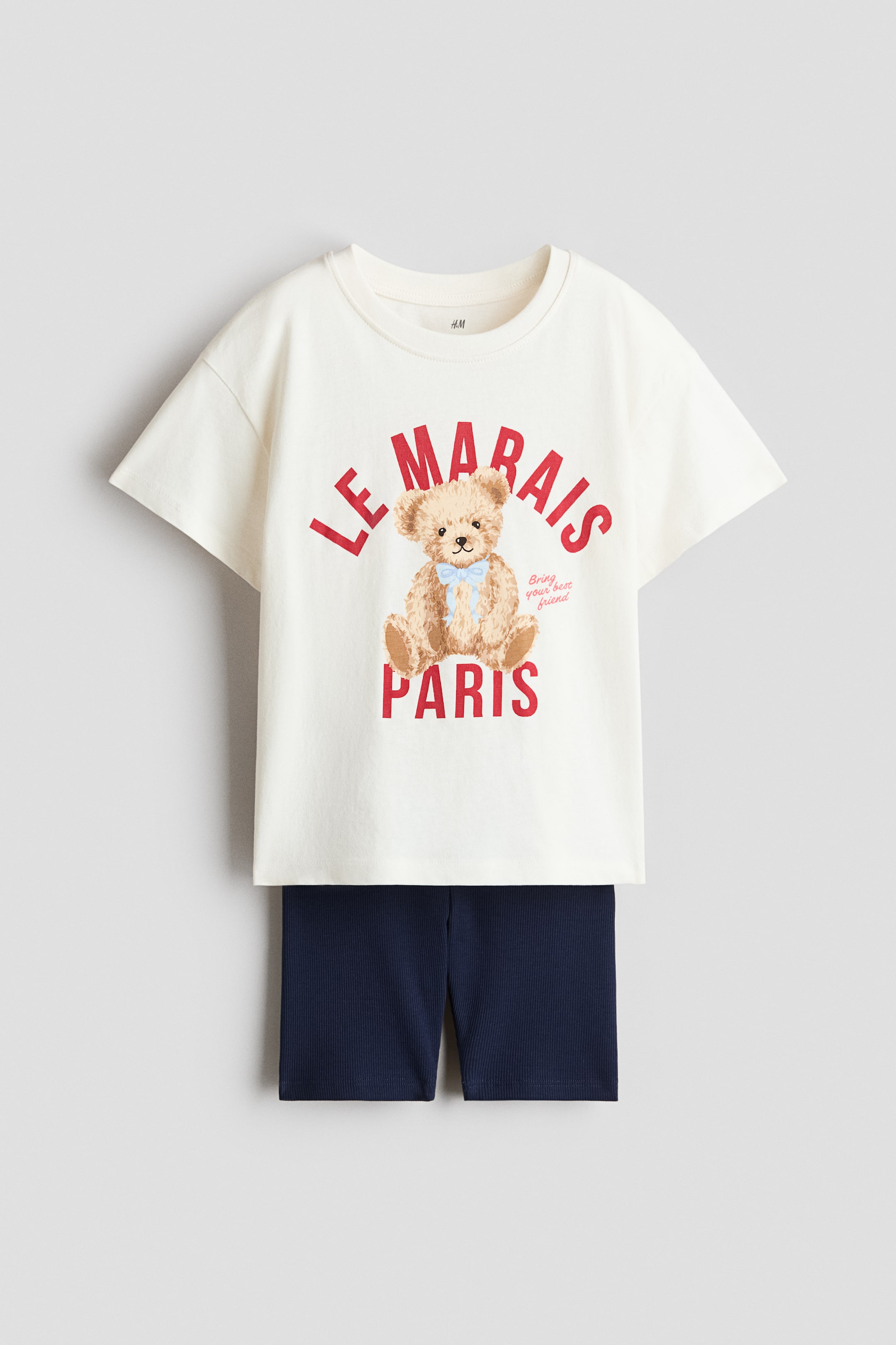 View larger image: A front-facing set features an off-white T-shirt and dark blue cycling shorts, both crafted from cotton jersey. The T-shirt showcases a red "LE MARAIS PARIS" print with a teddy bear, complemented by a ribbed trim around the neckline and dropped shoulders. The cycling shorts exhibit a ribbed texture and a covered elasticated waist.