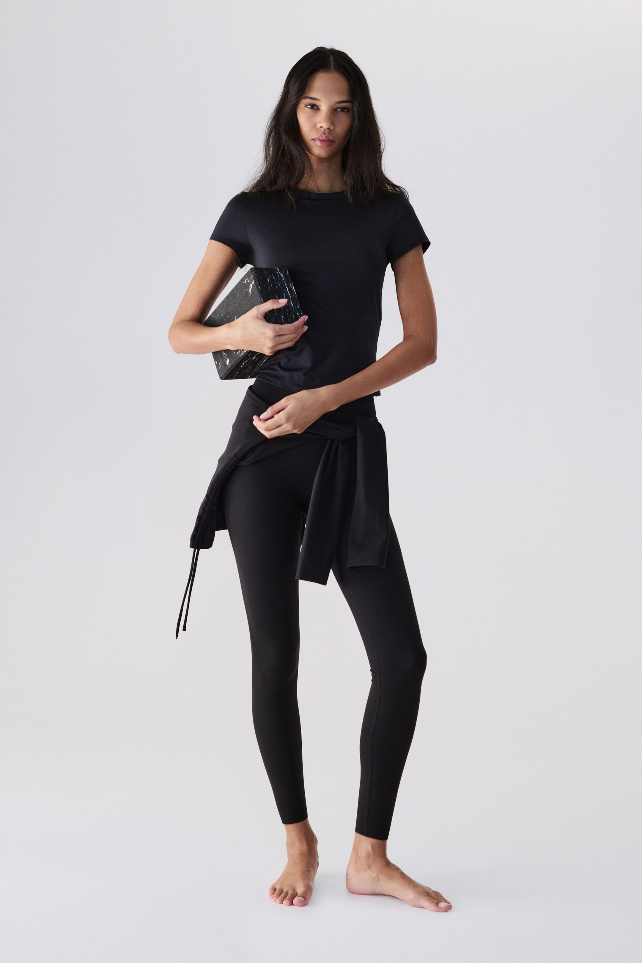 拡大画像を表示: 黒のフィットした半袖クルーネックトップが体にぴったりとフィットし、黒のフルレングスレギンスと組み合わせています。黒の長袖トップがウエストに結ばれ、袖が前面に巻きつき、ドローストリングが左側に垂れています。