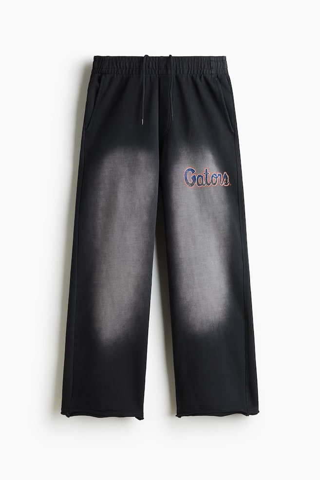 Black wide-leg sweatpants are shown from the front, featuring an elasticized waistband with a matching drawstring and side slip pockets. The pants display a prominent faded gray spray effect running vertically down the center of each leg, with a blue cursive "Gators" logo outlined in orange on the left thigh. - 1