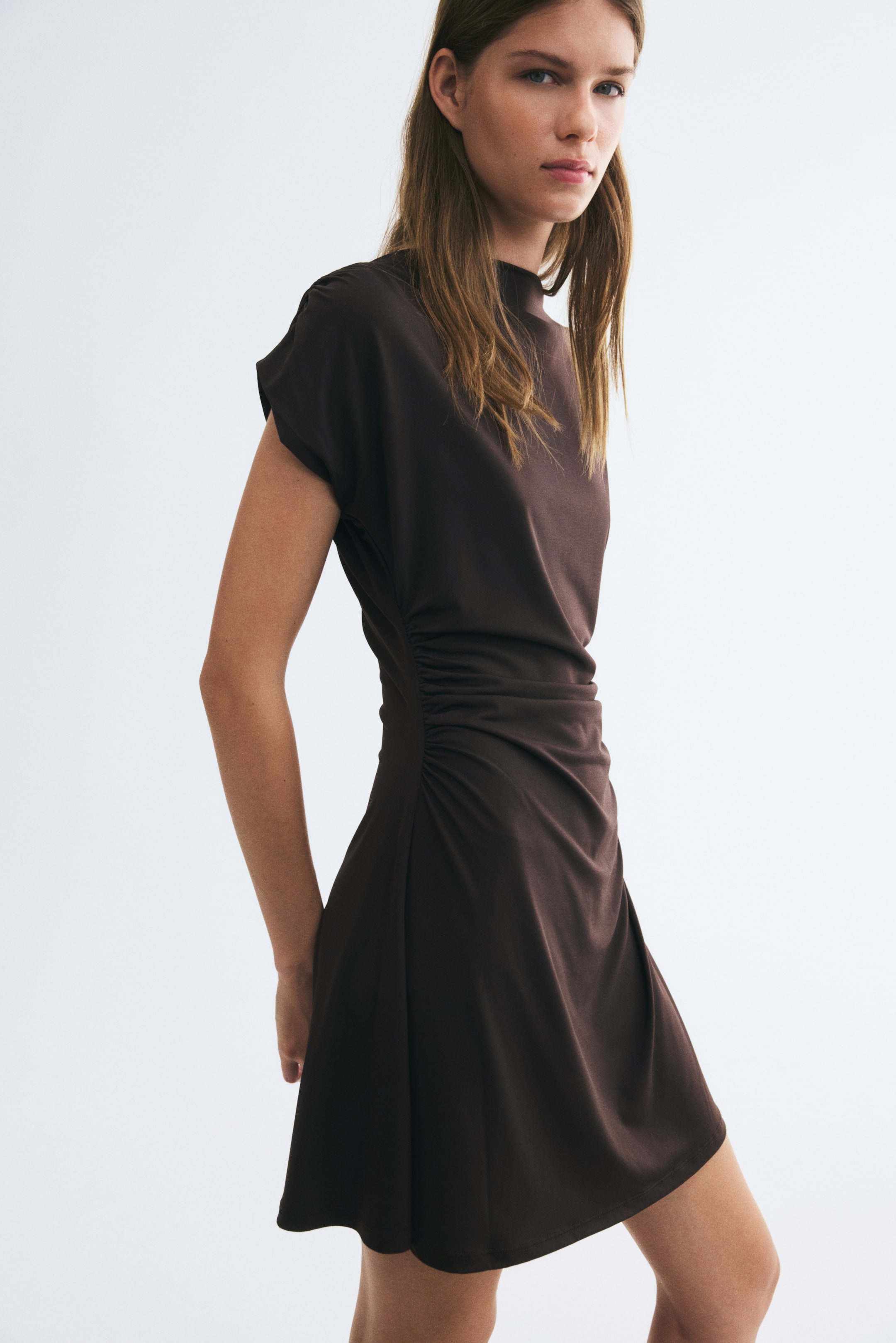 Ampliar la imagen: La modelo lleva puesto un vestido corto de jersey color café oscuro, con cuello barco alto, mangas tipo capa y costuras laterales fruncidas que crean un efecto drapeado sobre la cintura. El vestido se ajusta al cuerpo y presenta un dobladillo de corte recto.