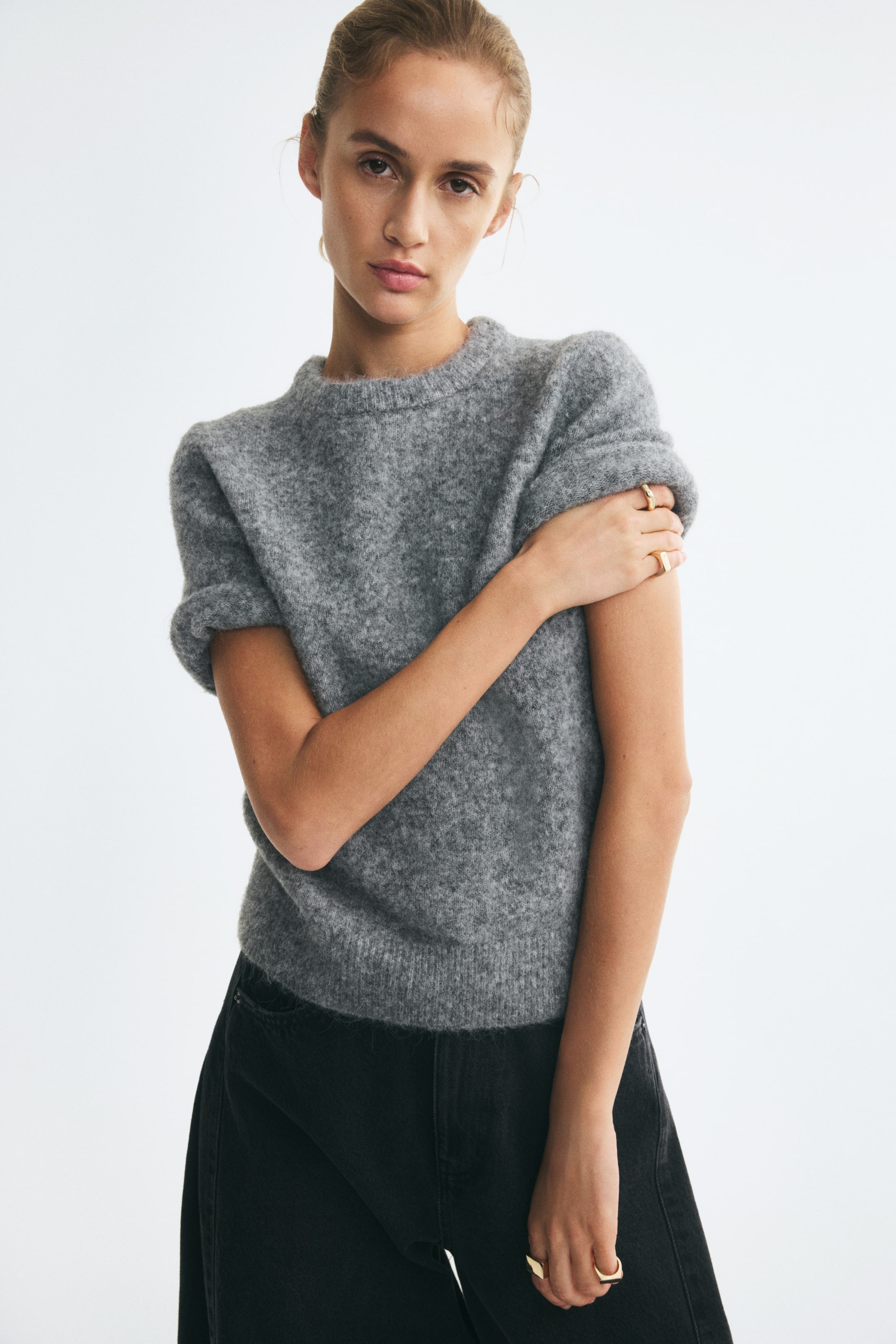 View larger image: The model is wearing a grey short-sleeve sweater in a soft, textured knit, holding one of the slightly rolled-up sleeves. The sweater features a round, rib-trimmed neckline and a ribbed hem, and is paired with black denim trousers.