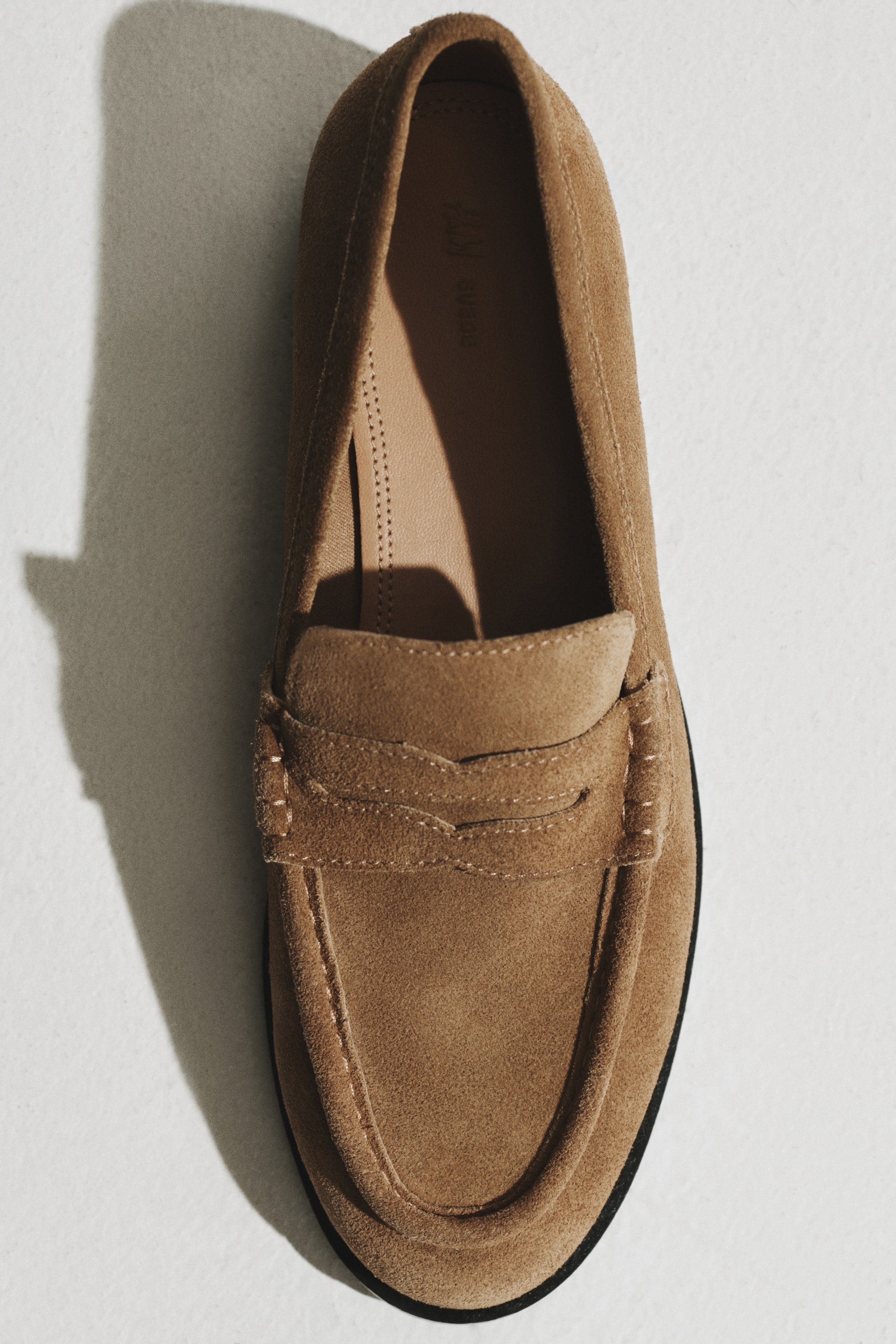 Visualizza immagine più grande: Un penny loafer in suede marrone chiaro è posizionato frontalmente, mostrando la costruzione moc-toe con cuciture ben visibili. Presenta la classica penny strap sulla vamp, una inner sole liscia color tan e una sottile suola nera che delinea il fondo.