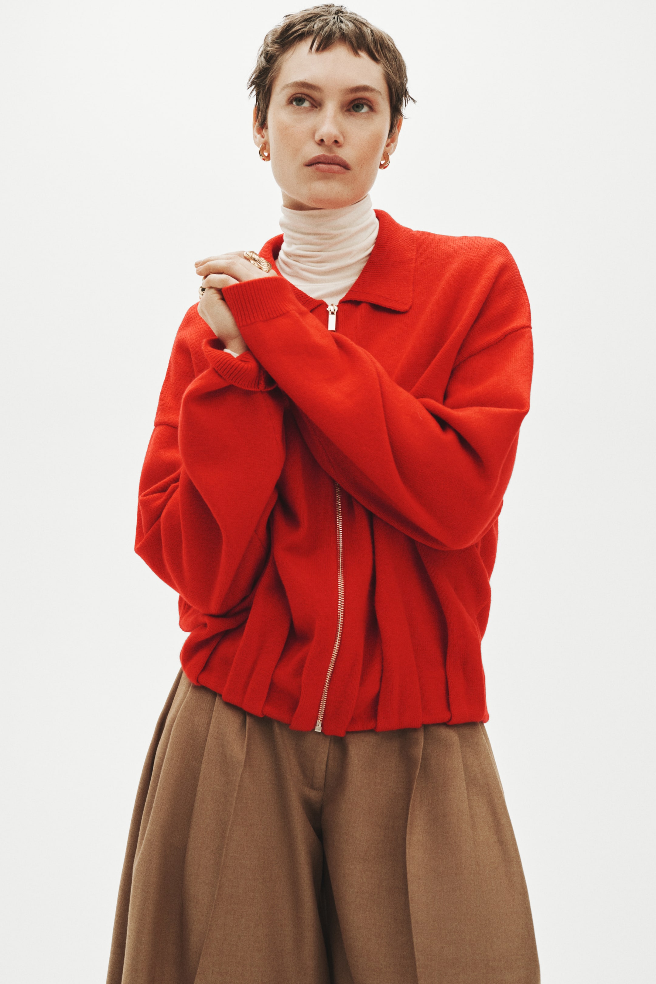 Xem ảnh lớn hơn: Chiếc áo khoác đan màu đỏ có cổ nhọn và khóa kéo mạ bạc dài suốt phía trước. Thiết kế với tay dài rộng rãi và gấu áo bo nhún, tạo phom blouson, được mặc ngoài áo cổ lọ màu kem.