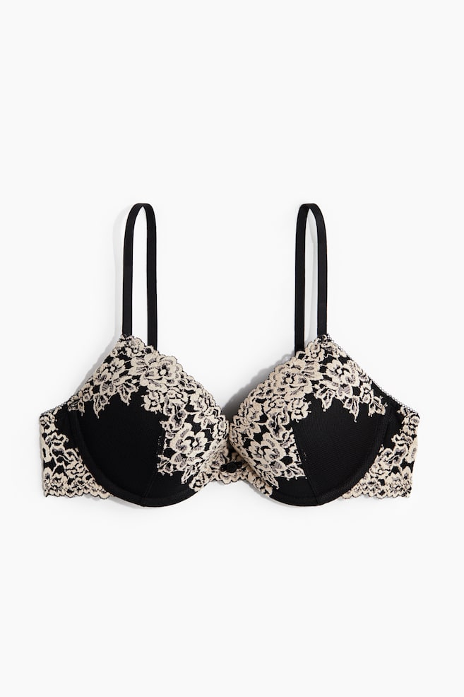 A front-facing bra is featuring black cups with an intricate beige floral lace overlay. The lace is covering the upper portion of the cups and extending along the underband, forming scalloped edges, while two black shoulder straps are rising from the top of the cups. - 1