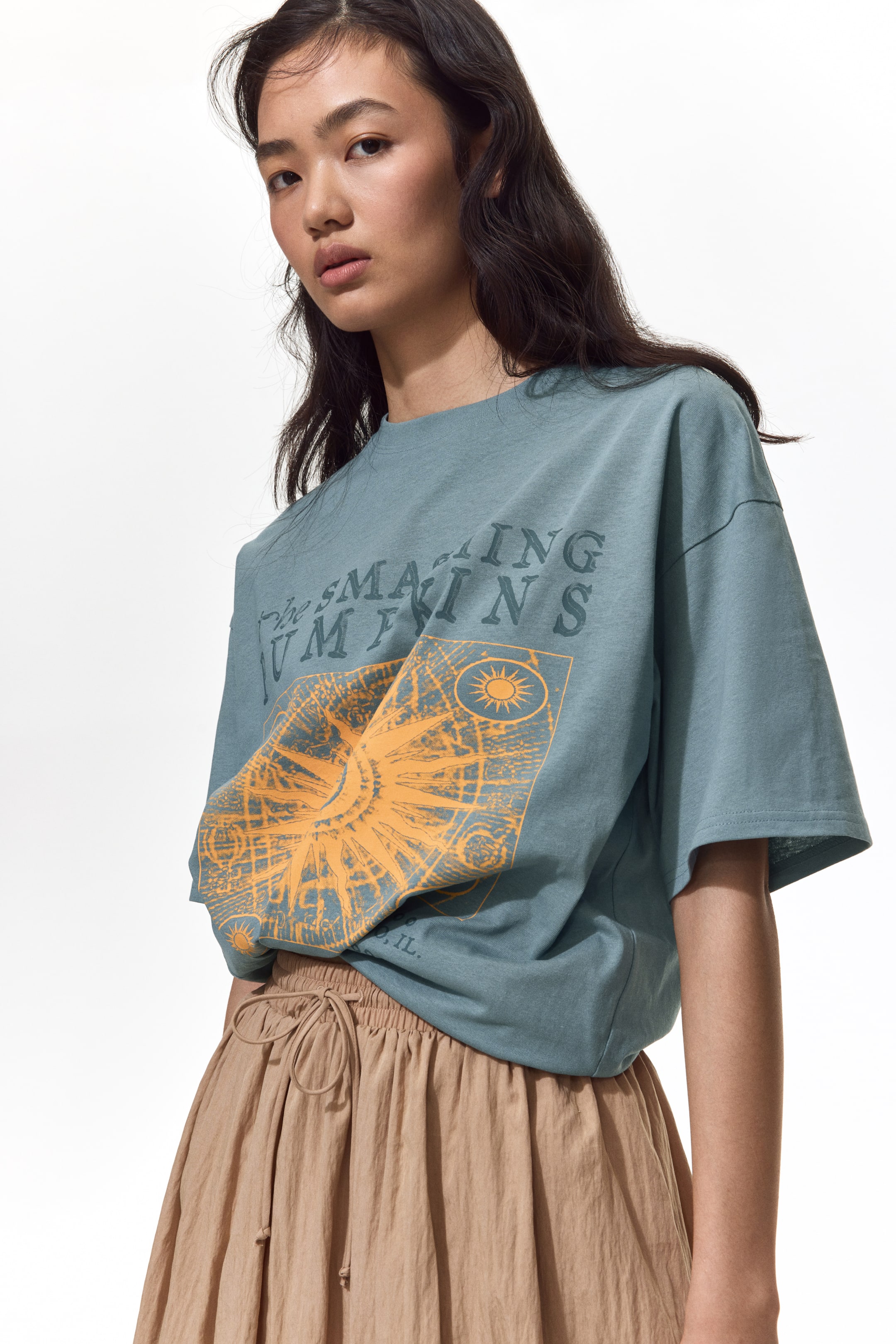 Agrandir l'image: Un t-shirt bleu sarcelle atténué présente un grand imprimé graphique orange délavé sur le devant, représentant un motif solaire avec des motifs astrologiques et un texte partiel. Il arbore une coupe oversize avec des épaules tombantes et des manches courtes, partiellement rentré à l'avant.