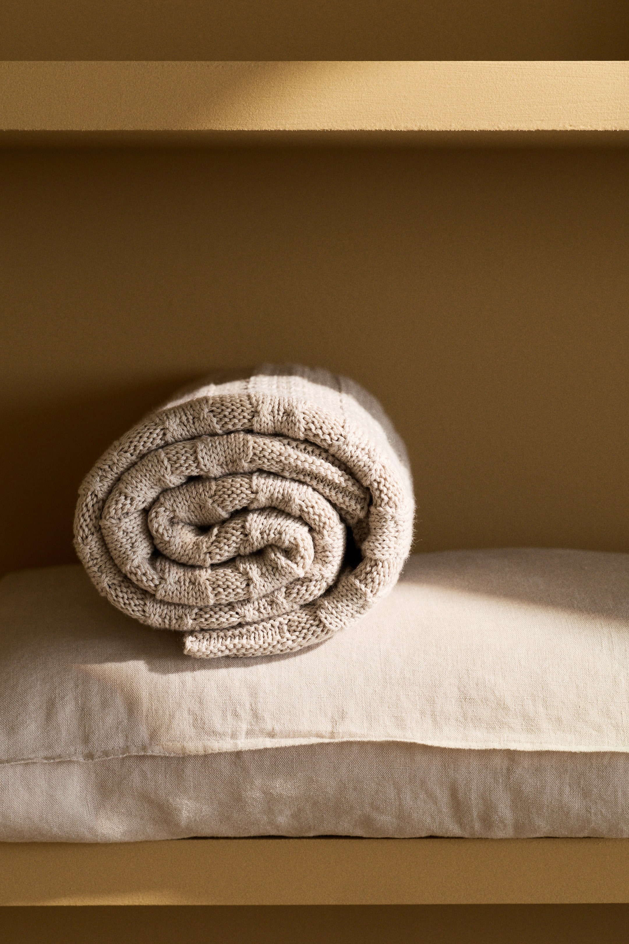 View larger image: A light beige knitted blanket is neatly rolled into a spiral, showcasing a basketweave knit pattern. It is placed on a light-coloured linen pillow.