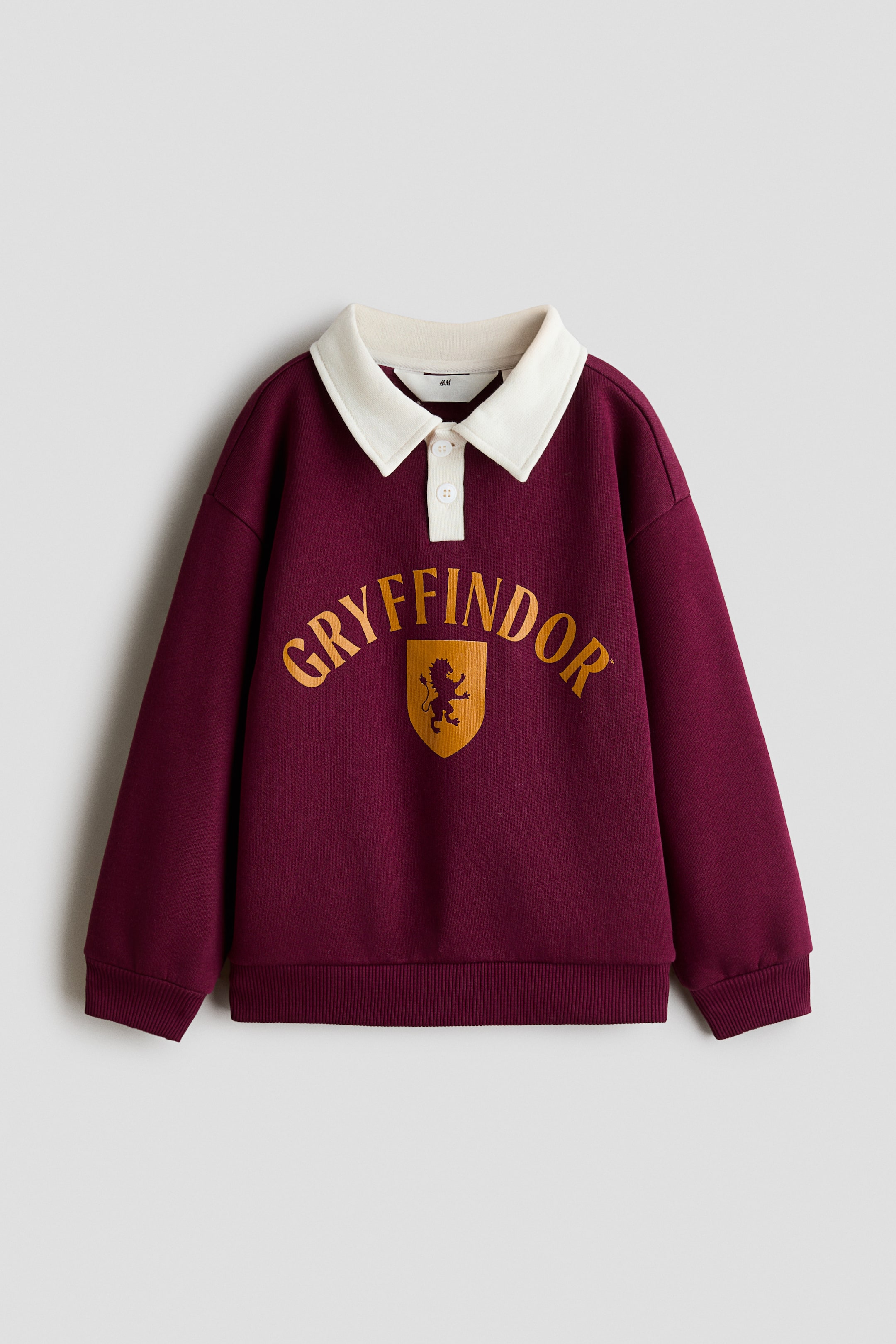 Grotere afbeelding bekijken: Een vooraanzicht van een bordeauxrode polo-sweatshirt dat plat ligt, met een contrasterende witte kraag en een sluiting met twee knopen. Op de borst staat de gebogen gouden tekst "GRYFFINDOR" boven een bijpassend gouden schildembleem met een leeuwensilhouet, en het kledingstuk is afgewerkt met geribde manchetten en een geribde zoom.