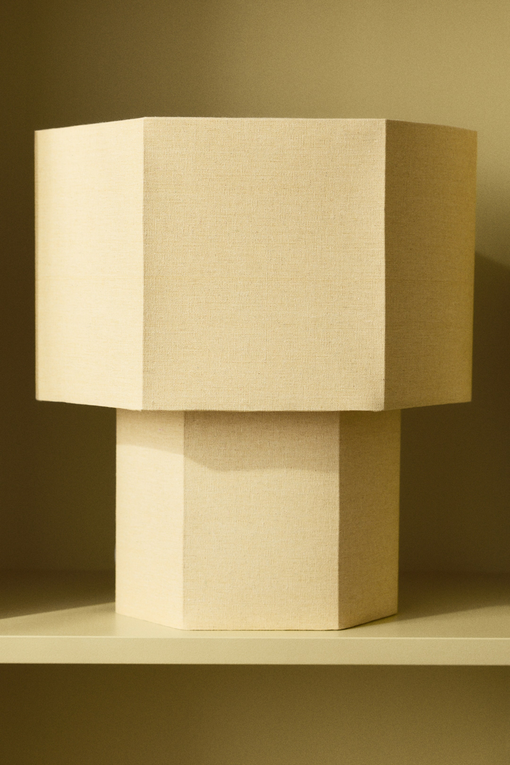 View larger image: A hexagonal lamp is showcasing a textured, light beige fabric shade and a matching hexagonal base. The broader shade sits atop the narrower base, with the lamp angled slightly to the right, highlighting its multi-faceted design.