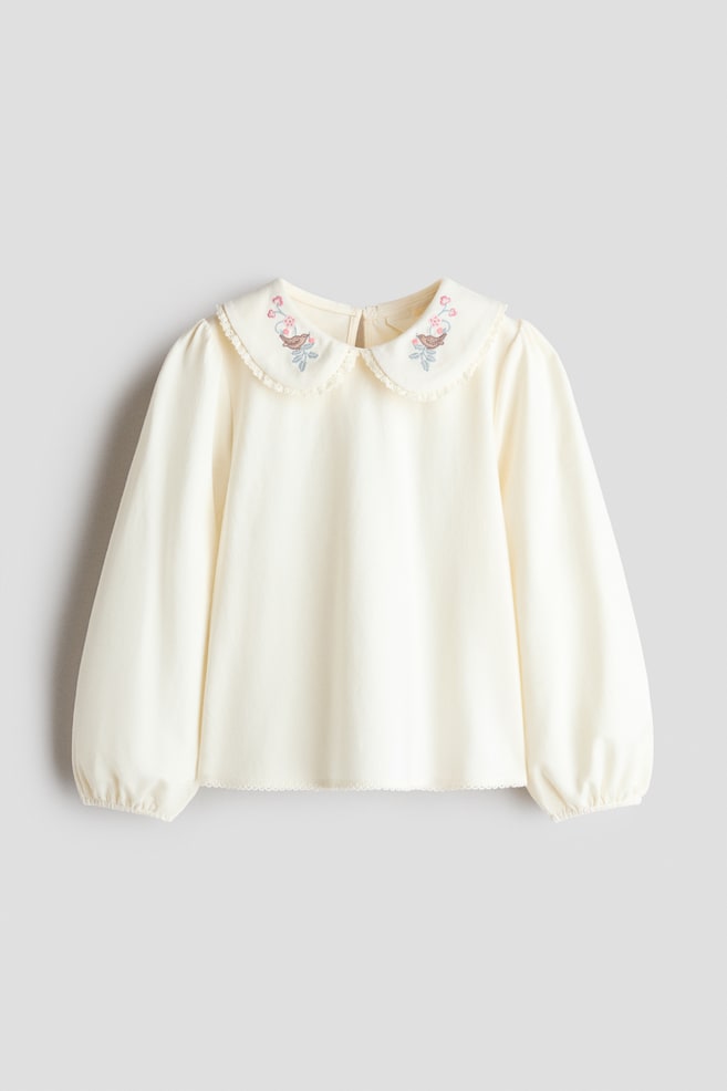 A cream-coloured cotton jersey blouse is shown from the front, featuring a rounded, frill-trimmed collar detailed with small embroidered flowers and birds. The blouse has long balloon sleeves with narrow, elasticated cuffs and a subtly scalloped hem. - 1