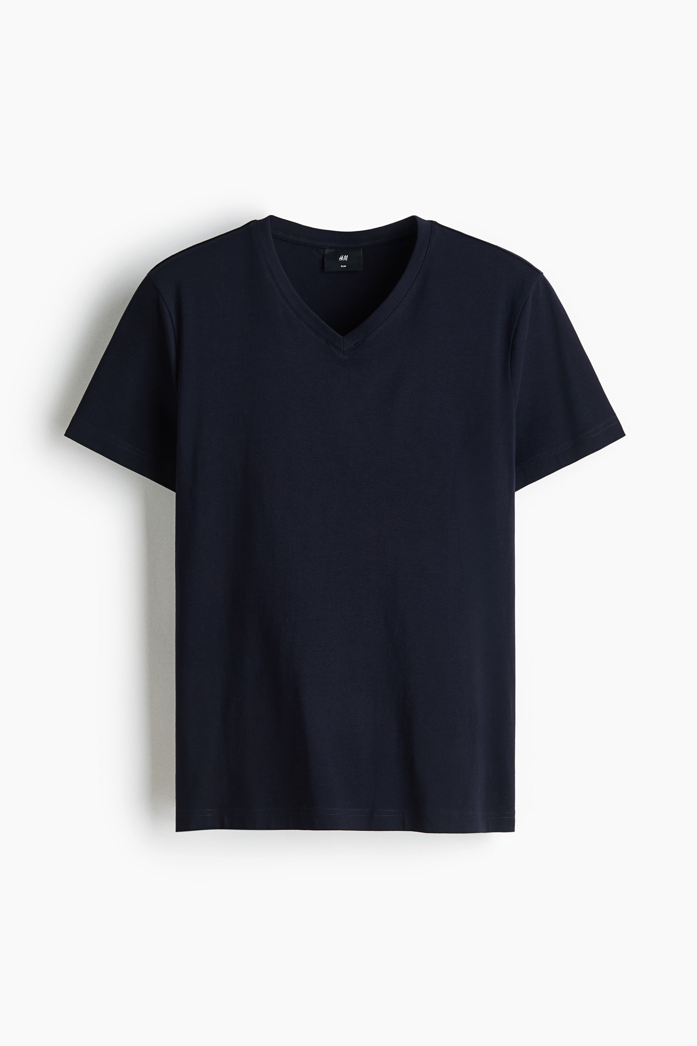 A dark navy blue T-shirt in a lightweight jersey fabric is worn by the model, featuring a slim fit, a rib-trimmed V-neck, and short sleeves. The T-shirt is paired with light cream trousers, with the model resting their hands in the trouser pockets.