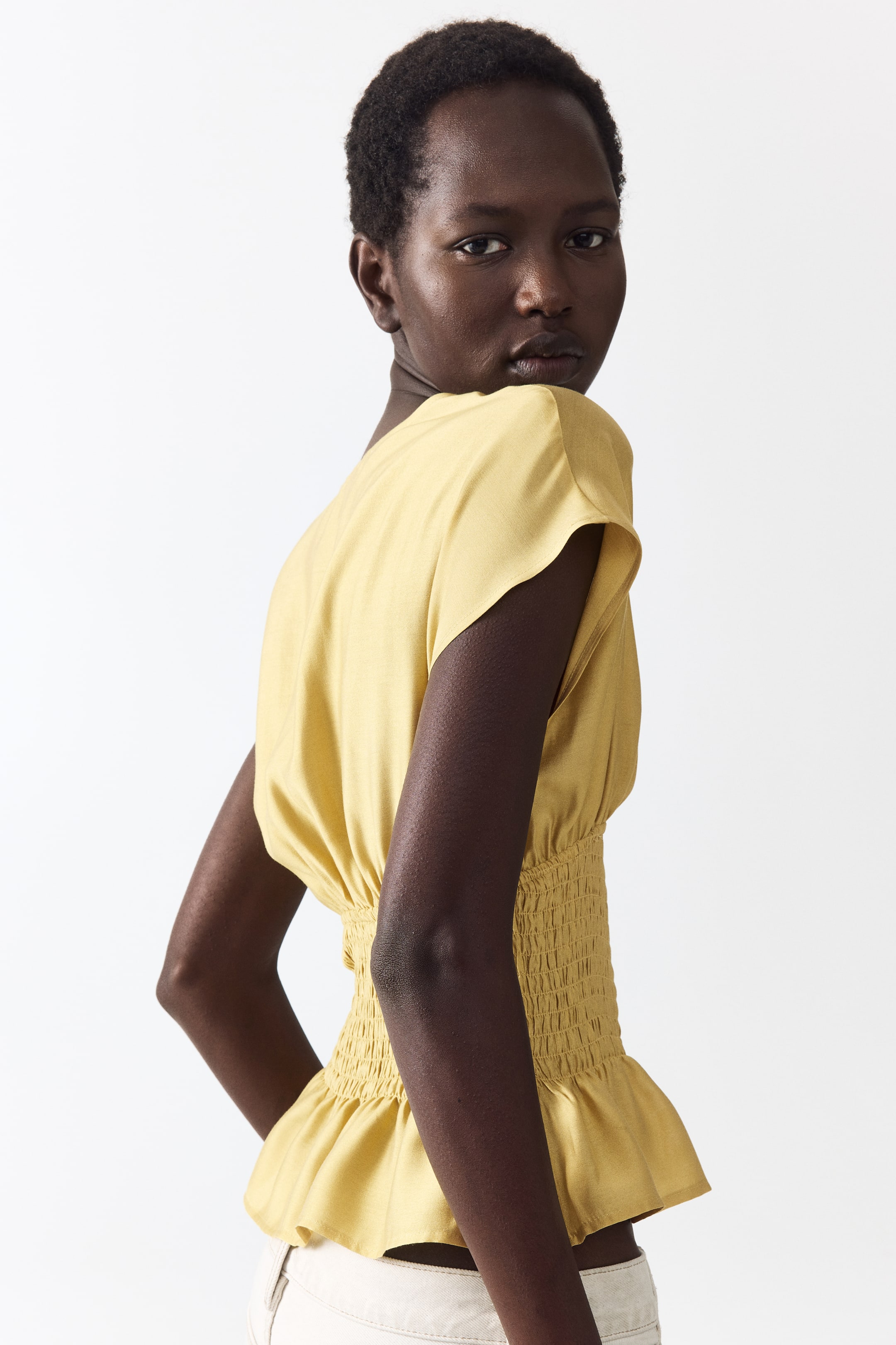 View larger image: A golden yellow top features short cap sleeves and a smocked, fitted waistline that cinches the garment, complemented by a peplum hem that flares out below. The top is showcased from a partial back view, emphasizing its structured waist.
