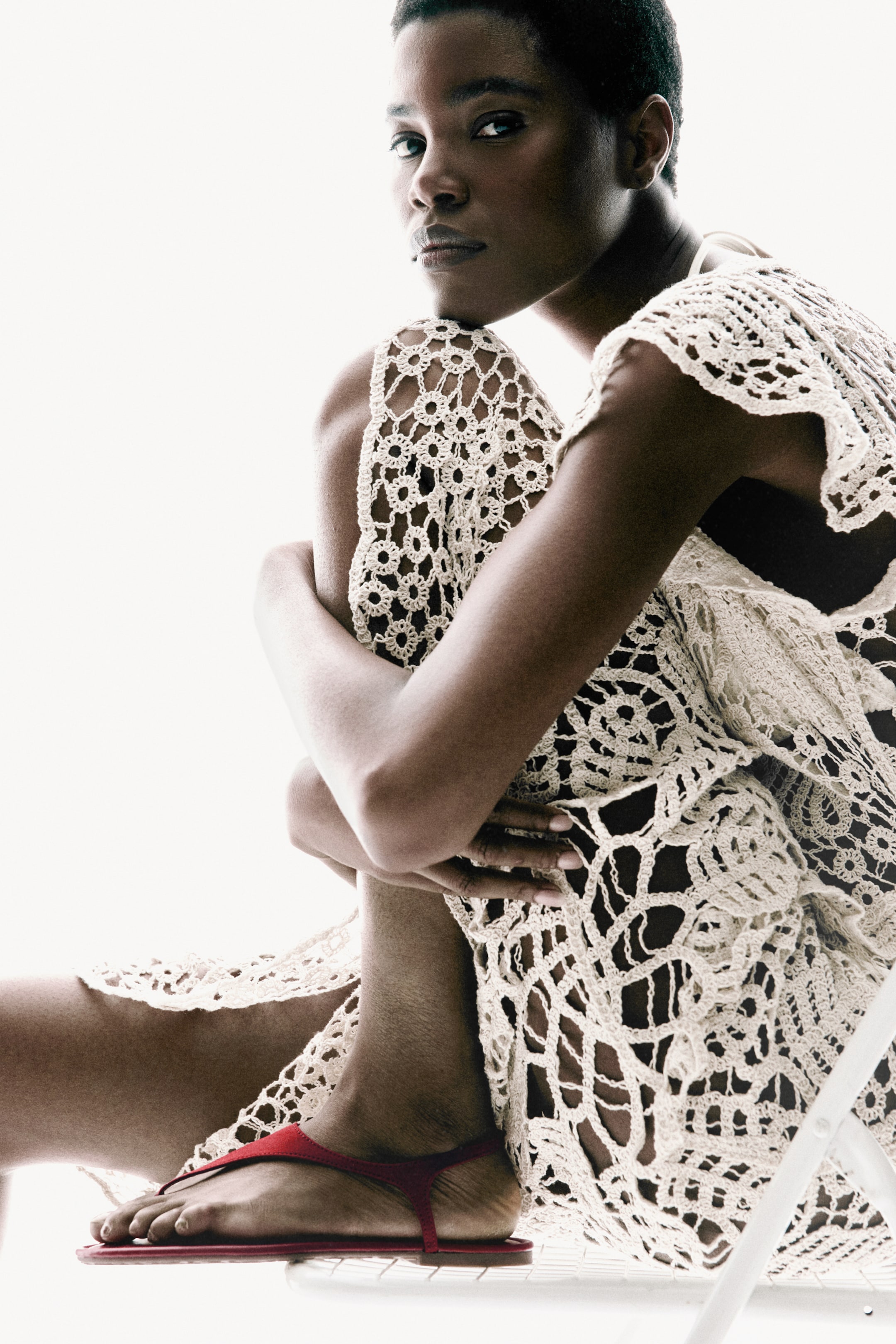 View larger image: A creamy off-white open-knit crocheted piece elegantly drapes over the shoulders and legs, showcasing intricate circular and geometric designs. On the left foot, a red flat thong sandal with slender straps and a flat sole is worn, captured from a side profile view.
